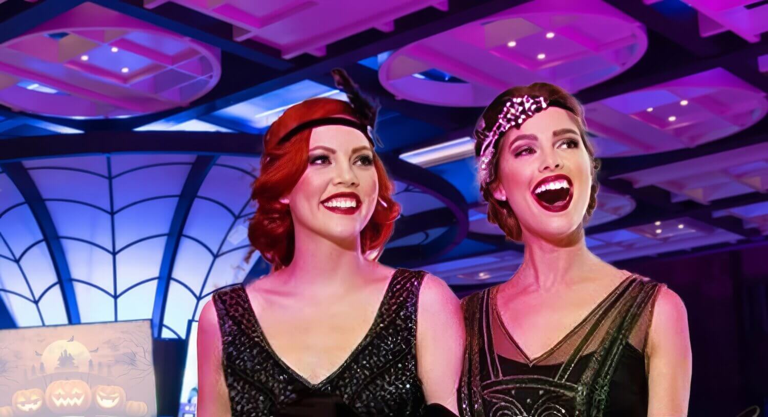 Two women in vintage 1920s flapper dresses and headbands smile and laugh at a festive event. The background features vibrant purple and blue lighting, a patterned ceiling, and a corner with a glowing Halloween-themed display including carved pumpkins.