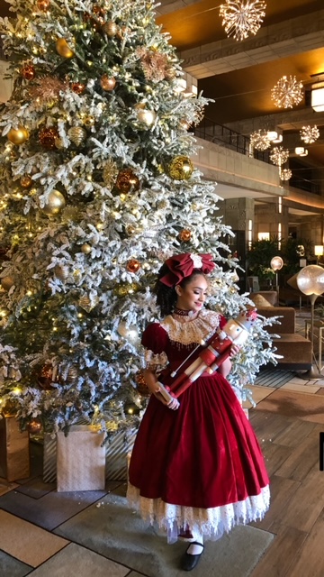 A child in a red velvet dress stands holding a nutcracker in front of a large, decorated Christmas tree with gold and white ornaments. The setting is a festive indoor space with elegant lighting and holiday decor.