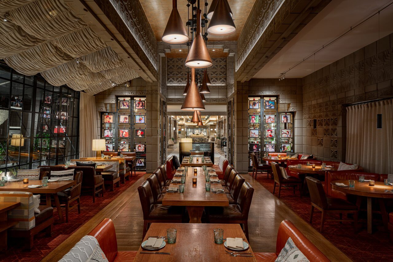 A warmly lit restaurant interior invites you to dine at wooden tables set for the evening. Copper pendant lights hang from the ceiling, casting a soft glow over a gallery wall adorned with colorful artwork. Cozy leather chairs line either side, offering comfort as guests savor their meals.