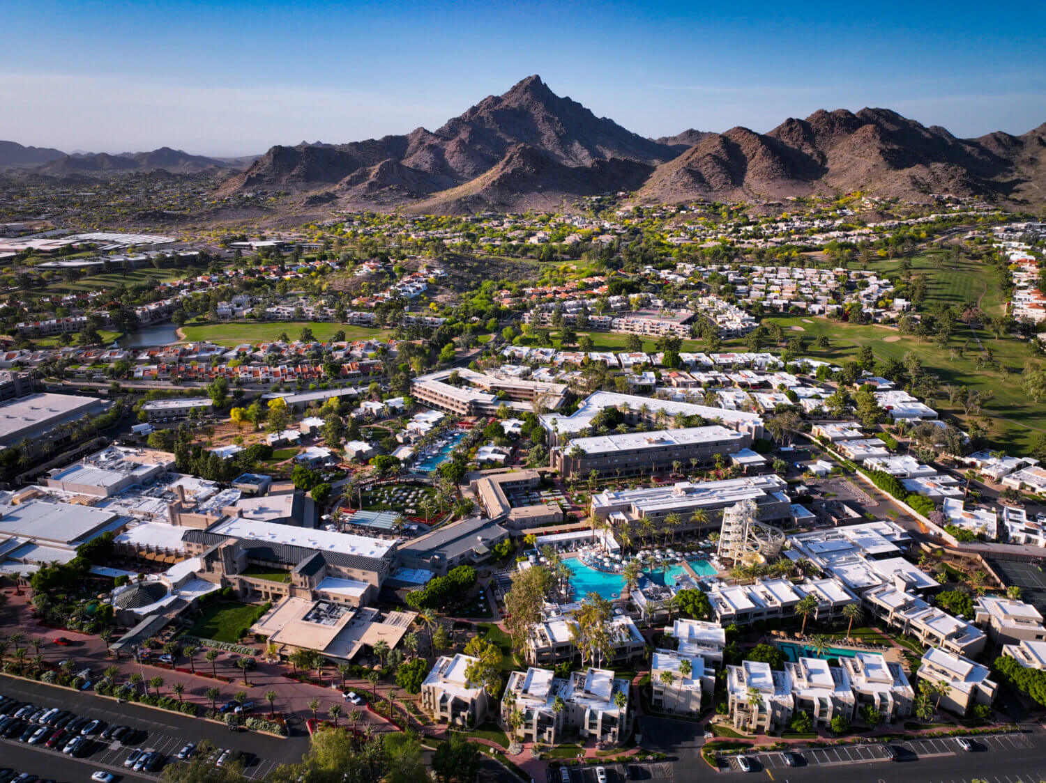 Aerial view of a resort surrounded by greenery and residential areas, featuring a large pool at the center and rugged mountains in the background under a clear blue sky—discover it with our exclusive Four Corner State Offer.