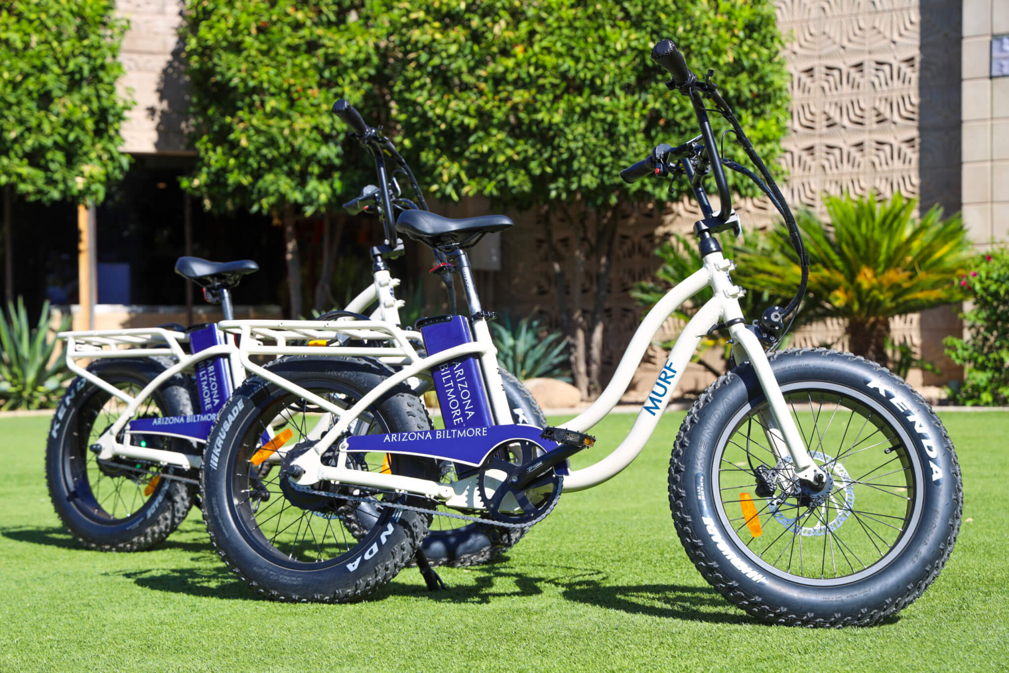 Two white electric bikes with wide tires are parked on green grass. The bikes have a blue panel labeled “Arizona Biltmore.” Trees and a building with patterned walls are in the background.