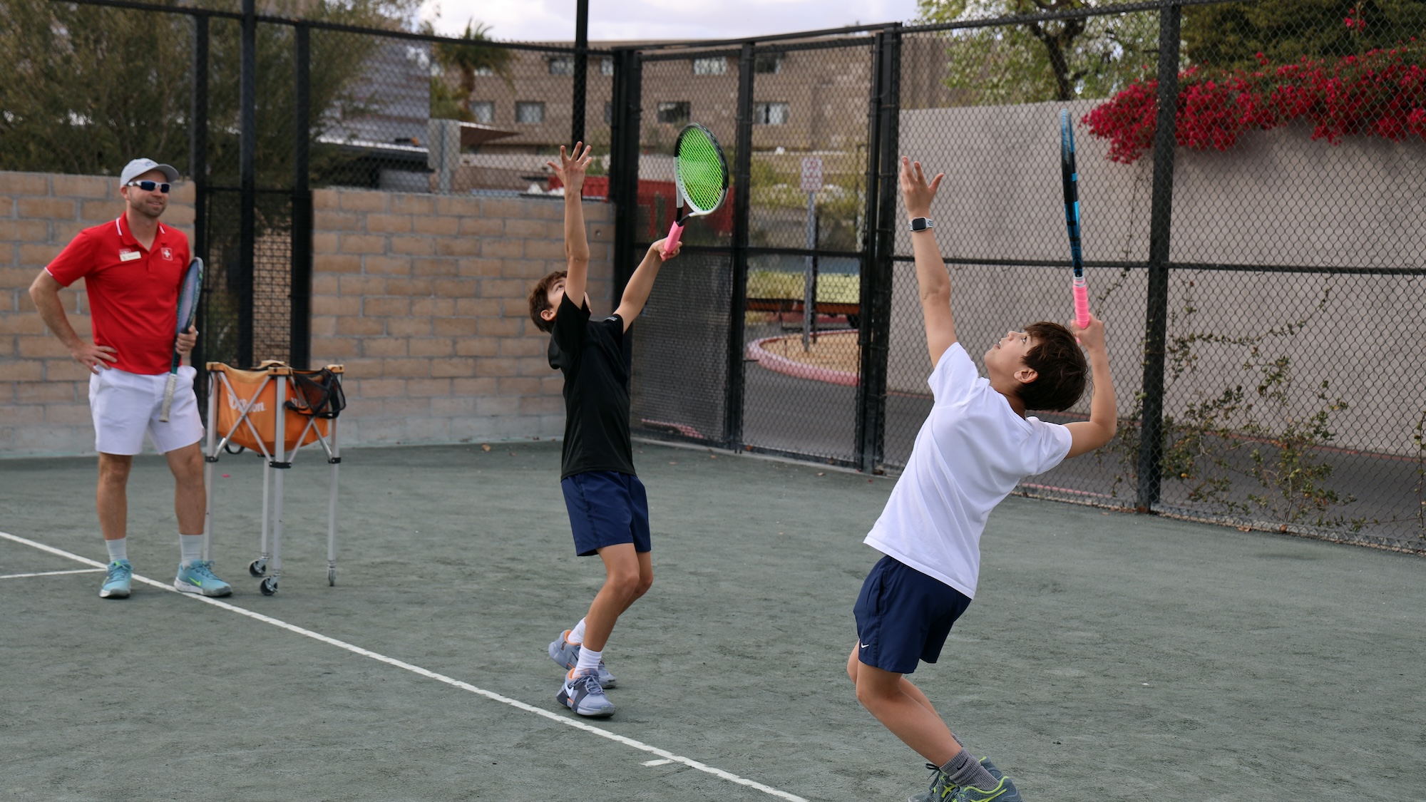 Two boys on a tennis court practice serving while a coach watches nearby. Both boys are mid-serve, holding tennis rackets and looking upward, with one wearing a black shirt and the other a white shirt.