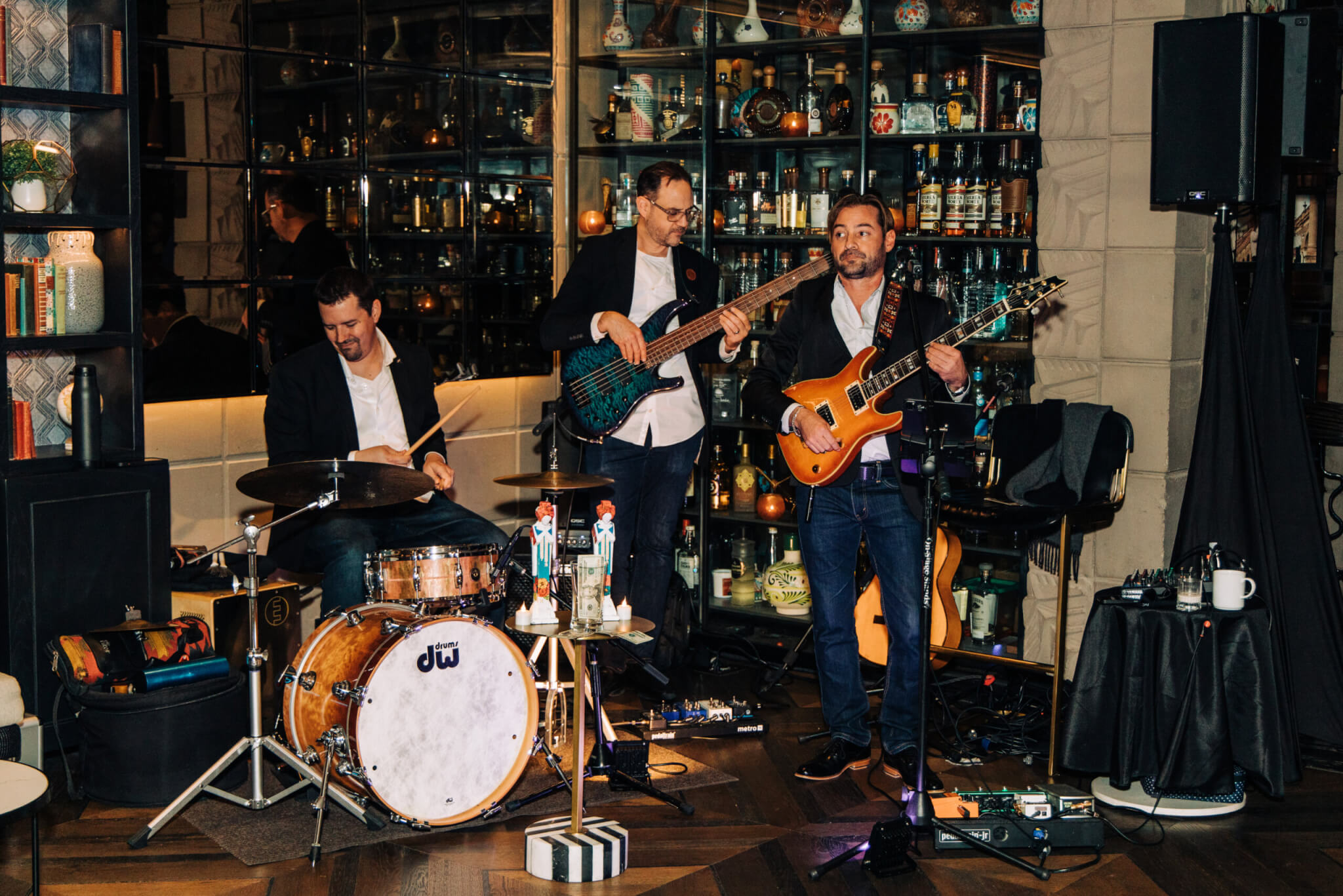 Three musicians perform indoors: one on drums, one playing bass guitar, and one playing electric guitar and singing. Shelves filled with bottles and various decor items are visible in the background.