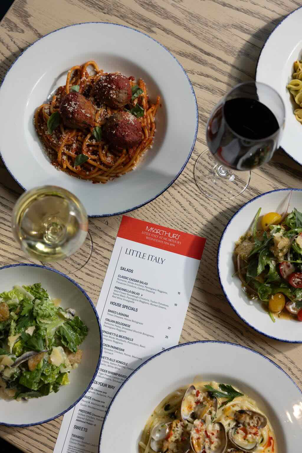 Overhead view of a table with plates of pasta, salad, a menu labeled "Little Italy," tacos, and glasses of red and white wine on a light wooden surface.