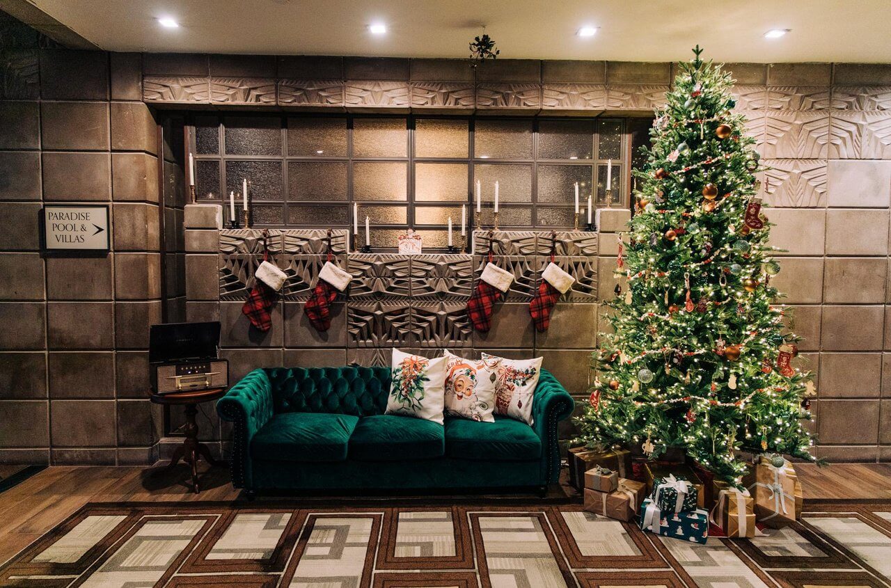 A decorated Christmas tree with lights and ornaments stands beside a green velvet sofa with festive pillows, bringing a touch of Christmas in July. Five stockings hang above the sofa on a patterned wall, with wrapped gifts resting under the tree.