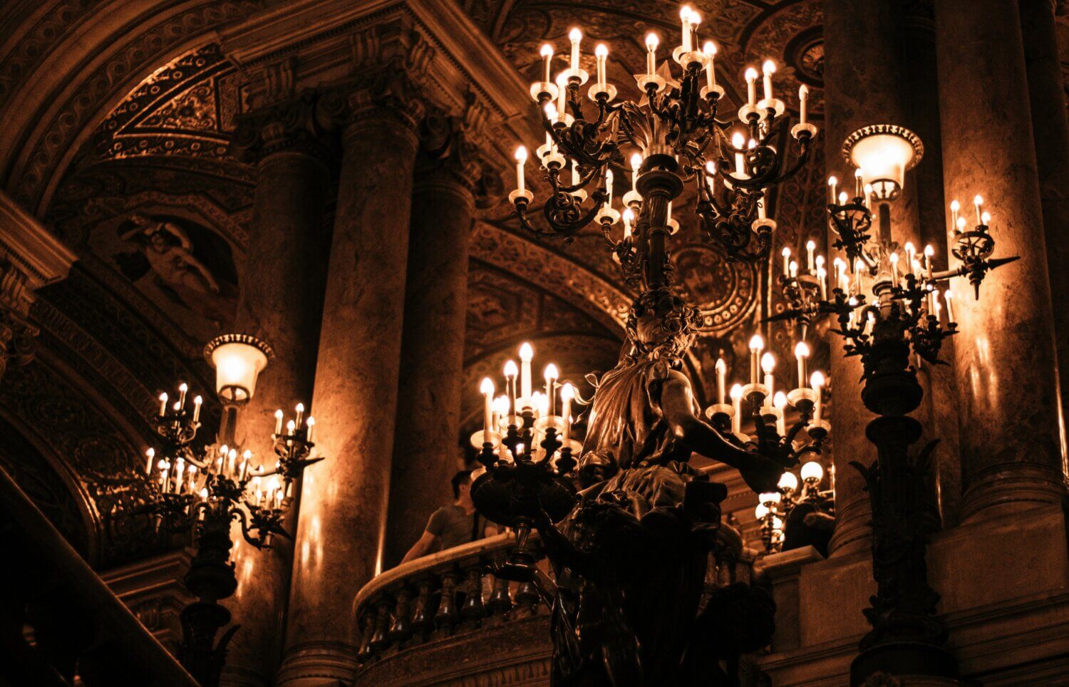 Ornate chandeliers and detailed ceiling decorations illuminate a grand hall with marble columns and a sculpted statue, creating a warm, elegant atmosphere perfect for a refined Halloween or Easter celebration.