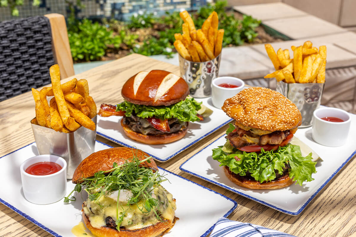 Three gourmet burgers with lettuce, tomato, cheese, and bacon are served on separate plates alongside golden French fries in metal cups and small containers of ketchup, all set on an outdoor wooden burger table.