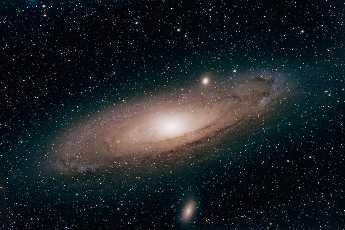 A detailed view of the Andromeda Galaxy, showing its bright, glowing center, spiral arms, and dust lanes, surrounded by countless stars and two smaller satellite galaxies in the background.