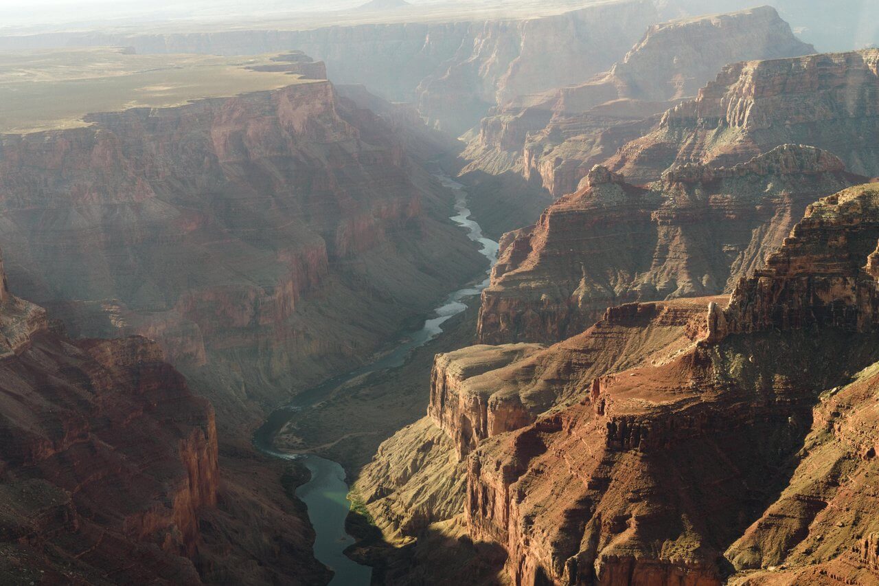 A sweeping aerial view of the Grand Canyon, as if seen during The Amethyst Expedition, with steep, layered red rock cliffs and a winding river coursing through the deep valley under a hazy sky.