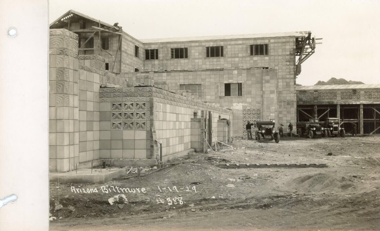Historic black and white photo of the Arizona Biltmore Hotel under construction in 1929, showing unfinished block walls, scaffolding, workers, vintage cars, and The Wonderbus parked near the building site.