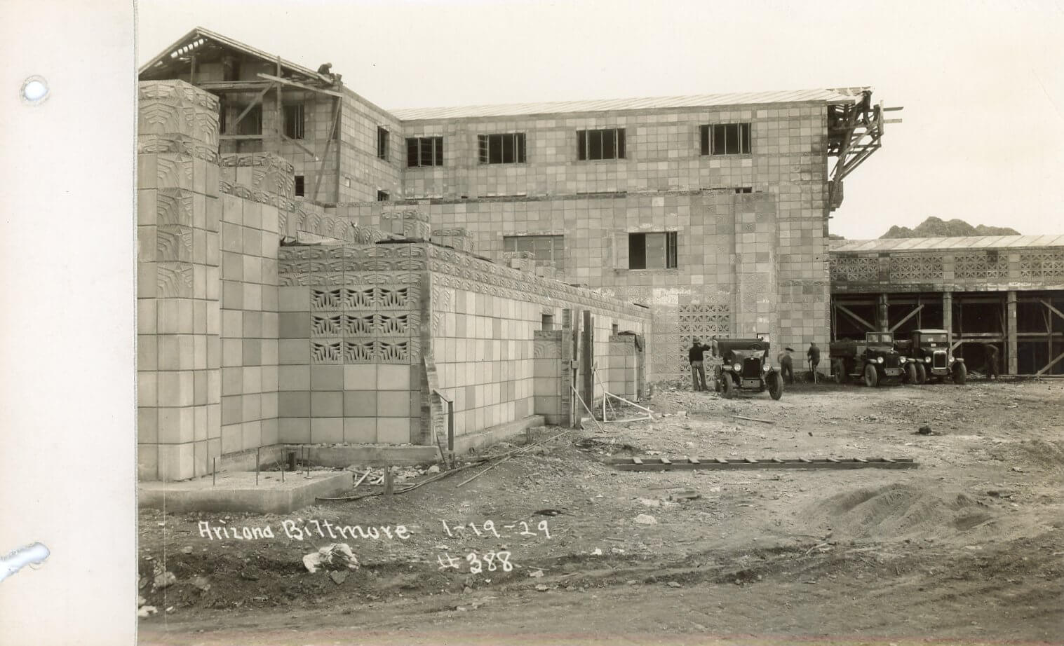Historic black and white photo of the Arizona Biltmore Hotel under construction in 1929, showing unfinished block walls, scaffolding, workers, vintage cars, and The Wonderbus parked near the building site.