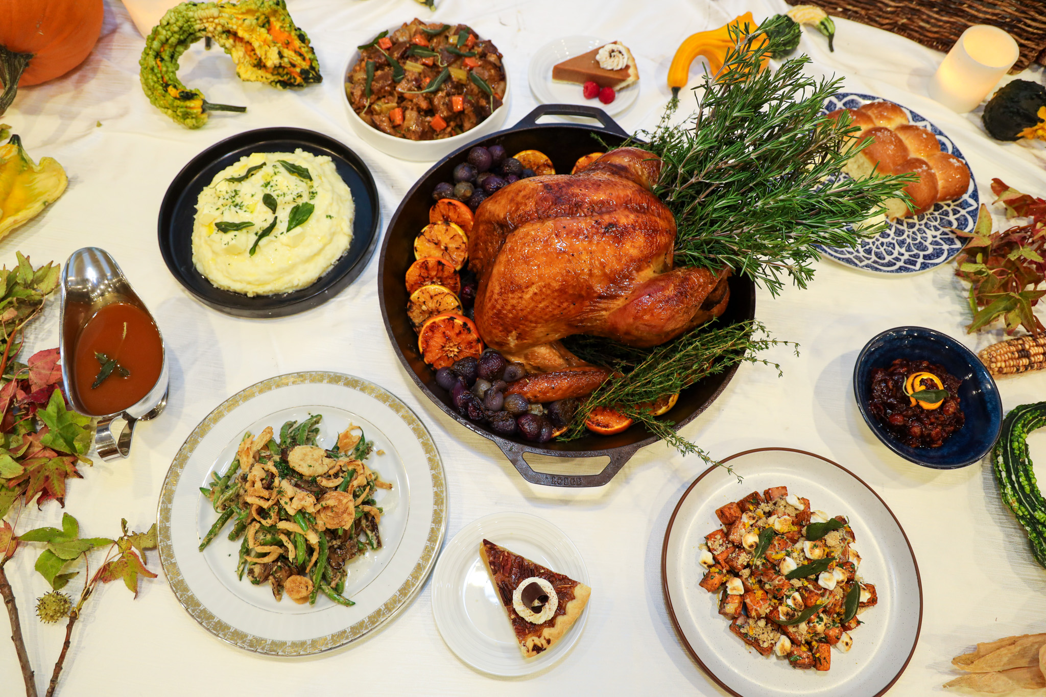 A Thanksgiving dinner spread featuring a roasted turkey with herbs, mashed potatoes, green bean casserole, stuffing, cranberry sauce, gravy, bread rolls, pie, and various autumn decorations on a white tablecloth.
