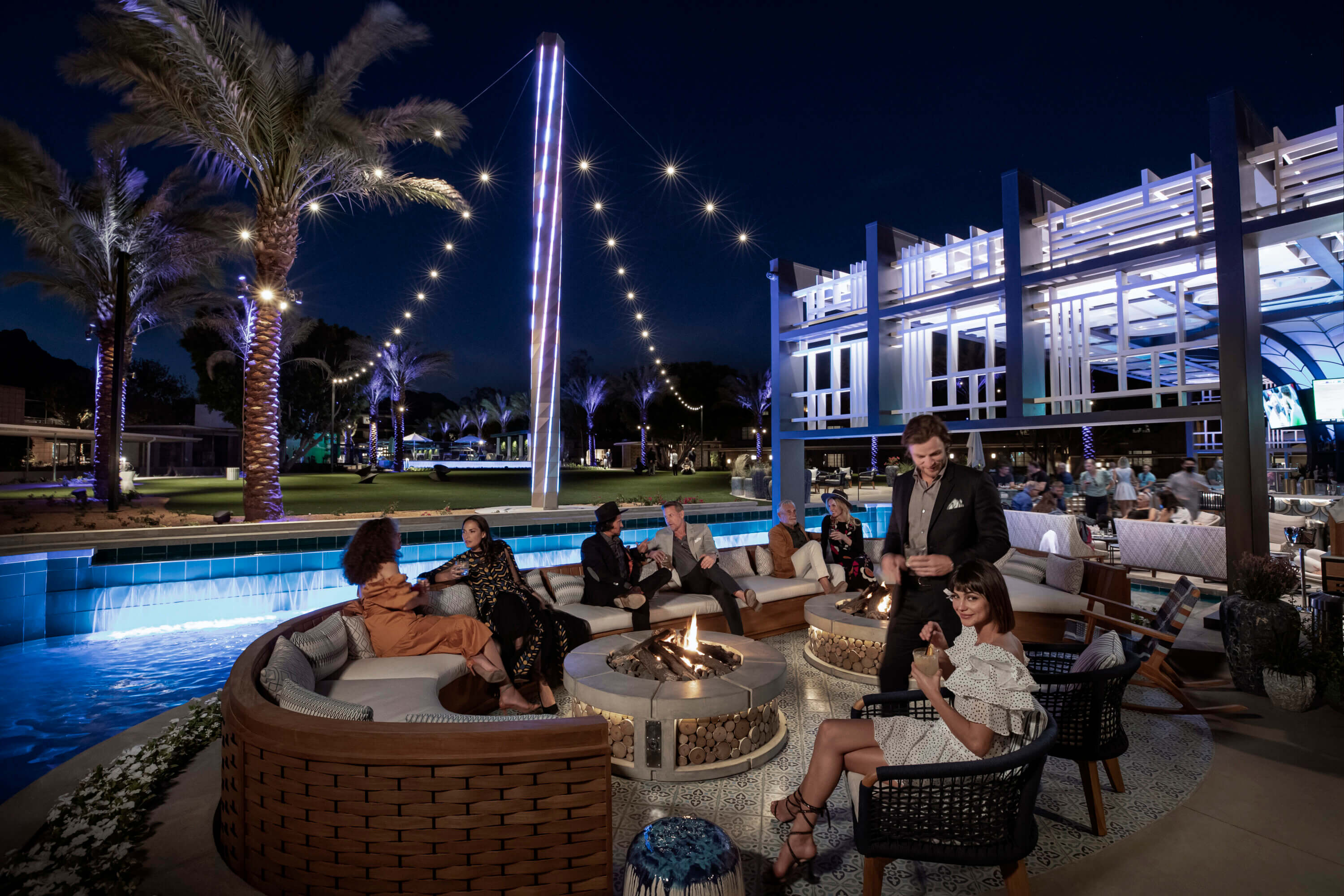 People sit around a circular fire pit at an outdoor lounge under palm trees and string lights. A server attends to guests as modern blue and white decor sets the scene for Your Championship Weekend Escape in a lively, upscale atmosphere.