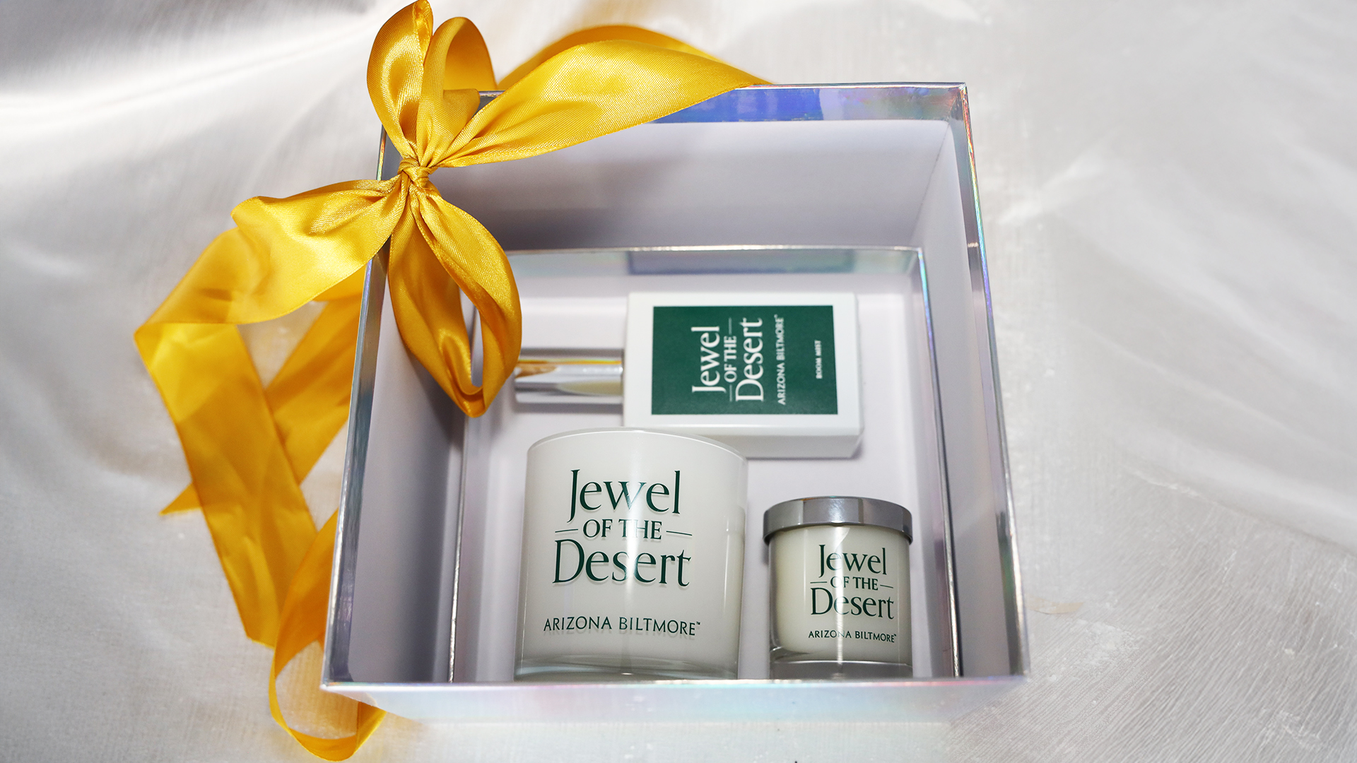 A white gift box with a yellow ribbon presents three "Jewel of the Desert" items—a candle, a small jar, and a spray bottle—on a silky white surface, making it a standout in The Ultimate Gift Guide.