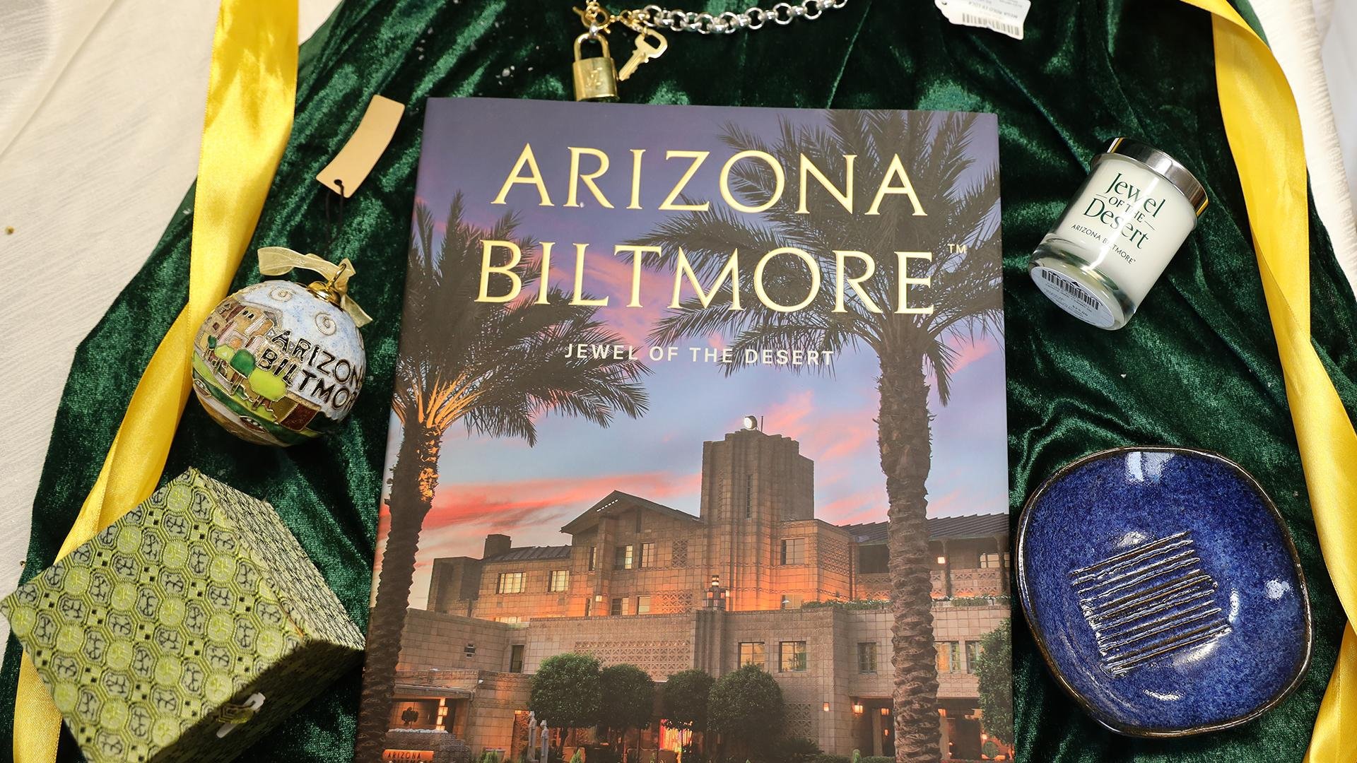 A book titled "Arizona Biltmore: Jewel of the Desert" lies on green velvet, surrounded by a Christmas ornament, small box, keychain, candle, and striped dish—an elegant scene for The Ultimate Gift Guide with a yellow ribbon draped alongside.