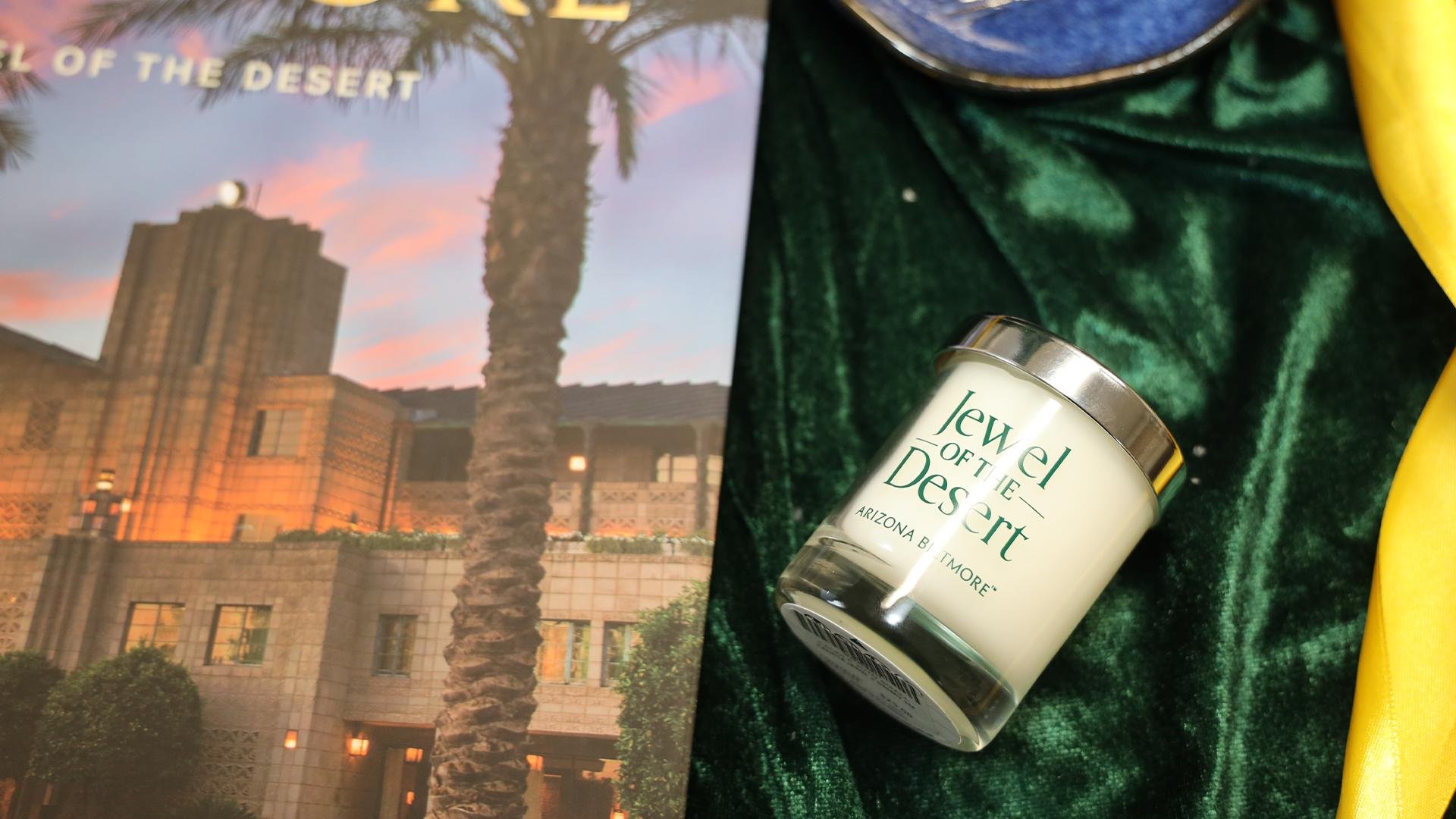 A candle labeled “Jewel of the Desert” sits on green velvet next to a book featuring The Wonderbus and a building with palm trees at sunset. A hint of yellow cloth and a blue dish complete this inviting scene.