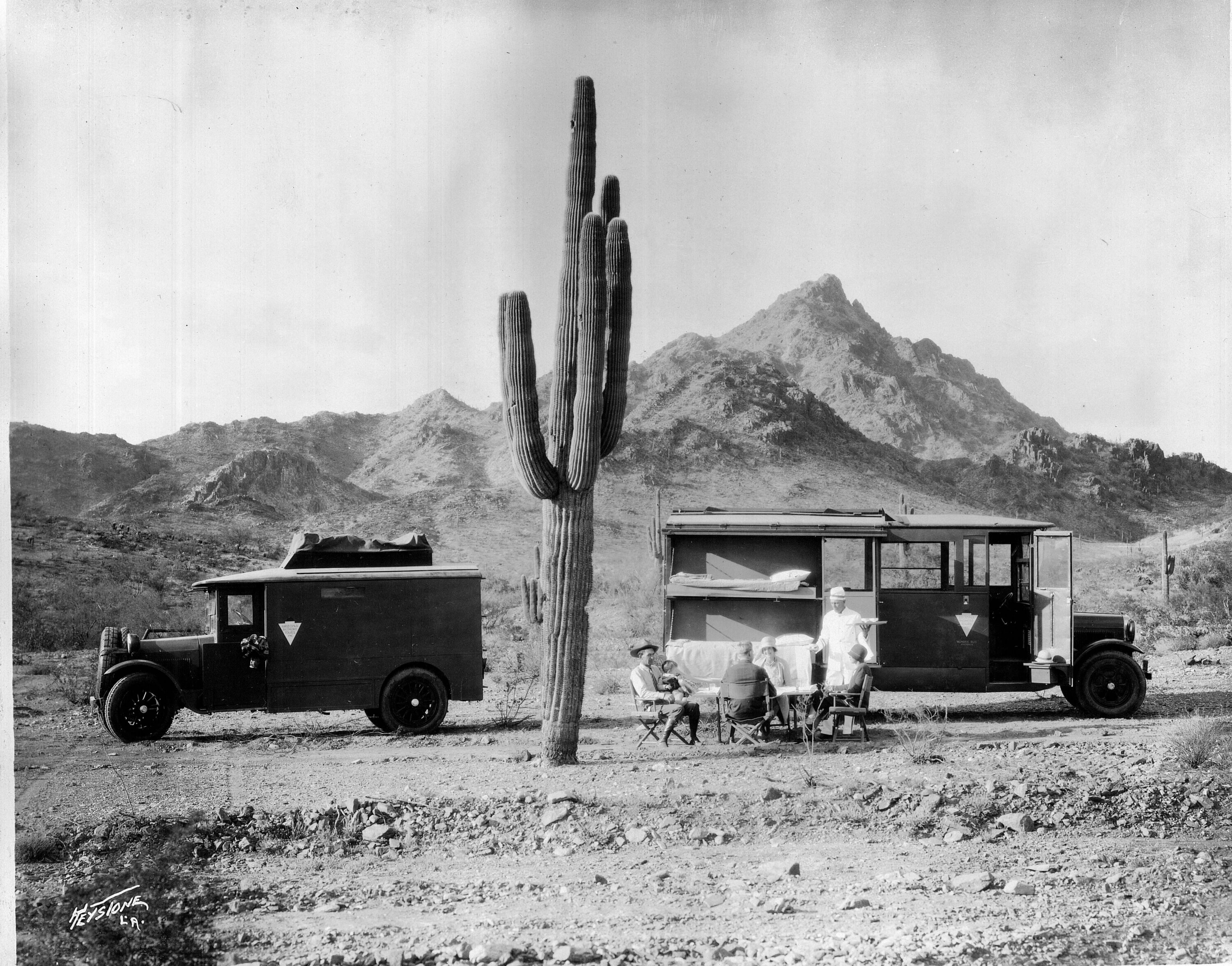 Two vintage camper vehicles, including The Wonderbus, are parked in a desert landscape with mountains behind them. A large saguaro cactus stands between them as four people sit at a table outside, enjoying a meal together.