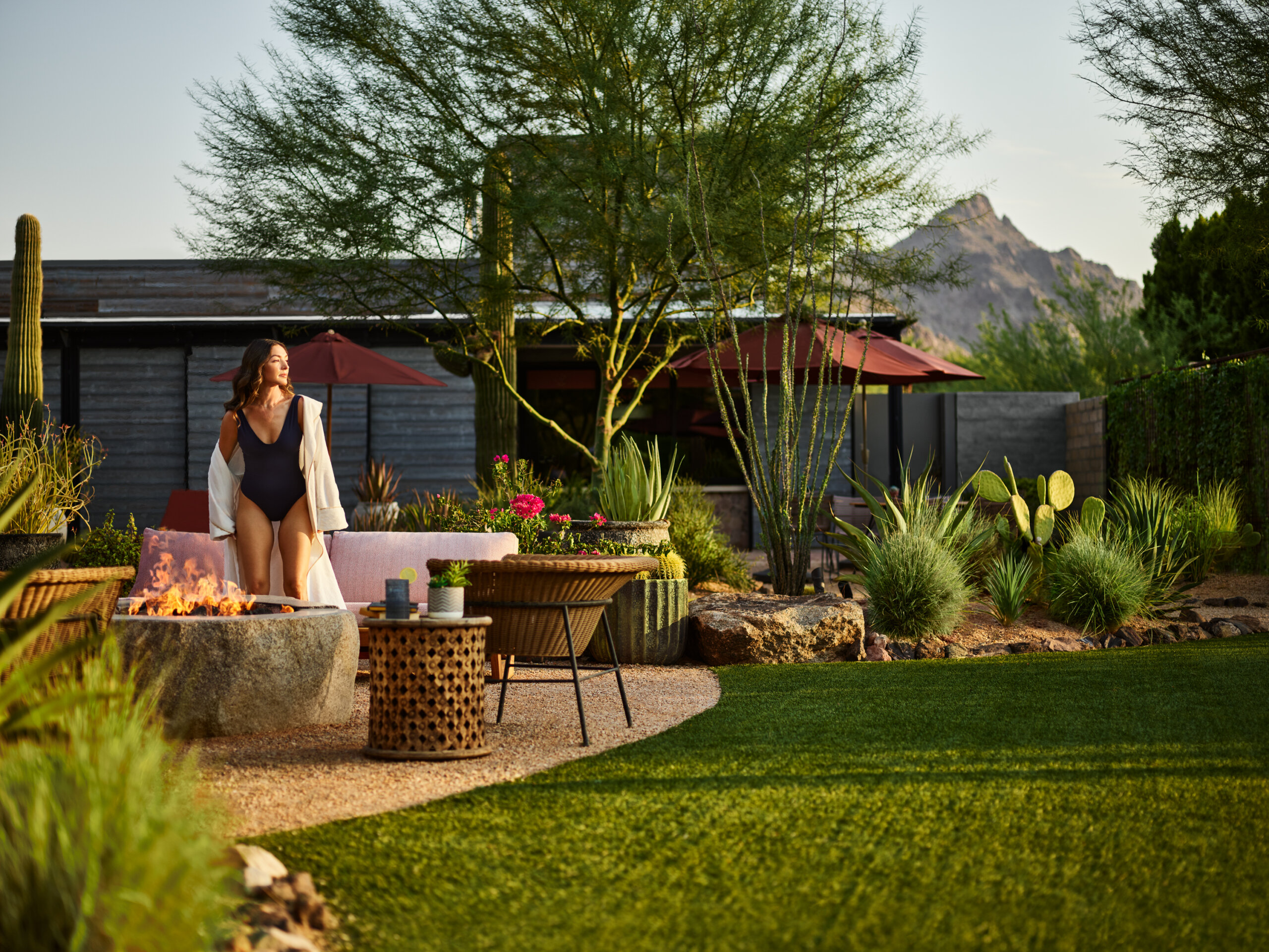 A woman in a black swimsuit and white cover-up stands by an outdoor fire pit surrounded by lush desert landscaping, patio furniture, and mountains in the background.