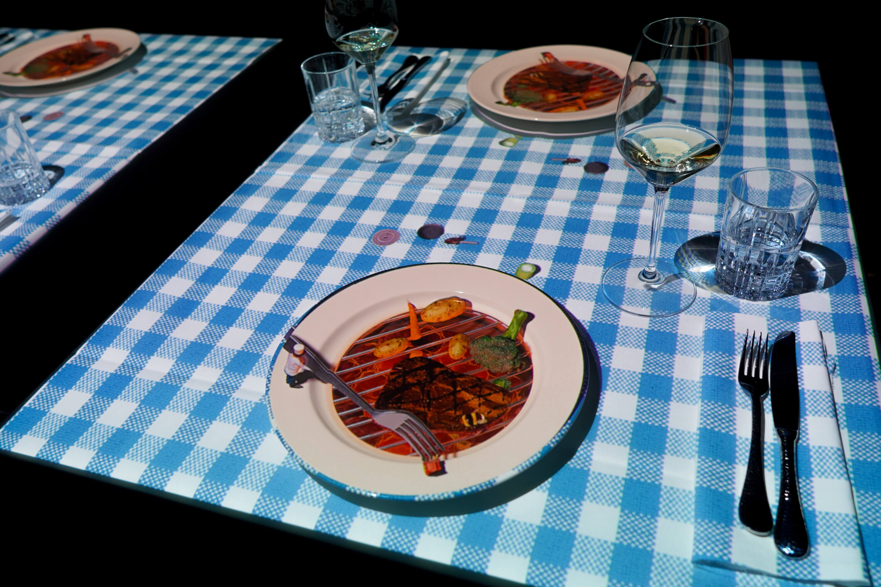 A plate with a digital projection of grilled food is set on a blue and white checkered tablecloth, surrounded by glasses, silverware, and a wine glass, capturing the Last Tastes of Summer in an illusion of a meal.