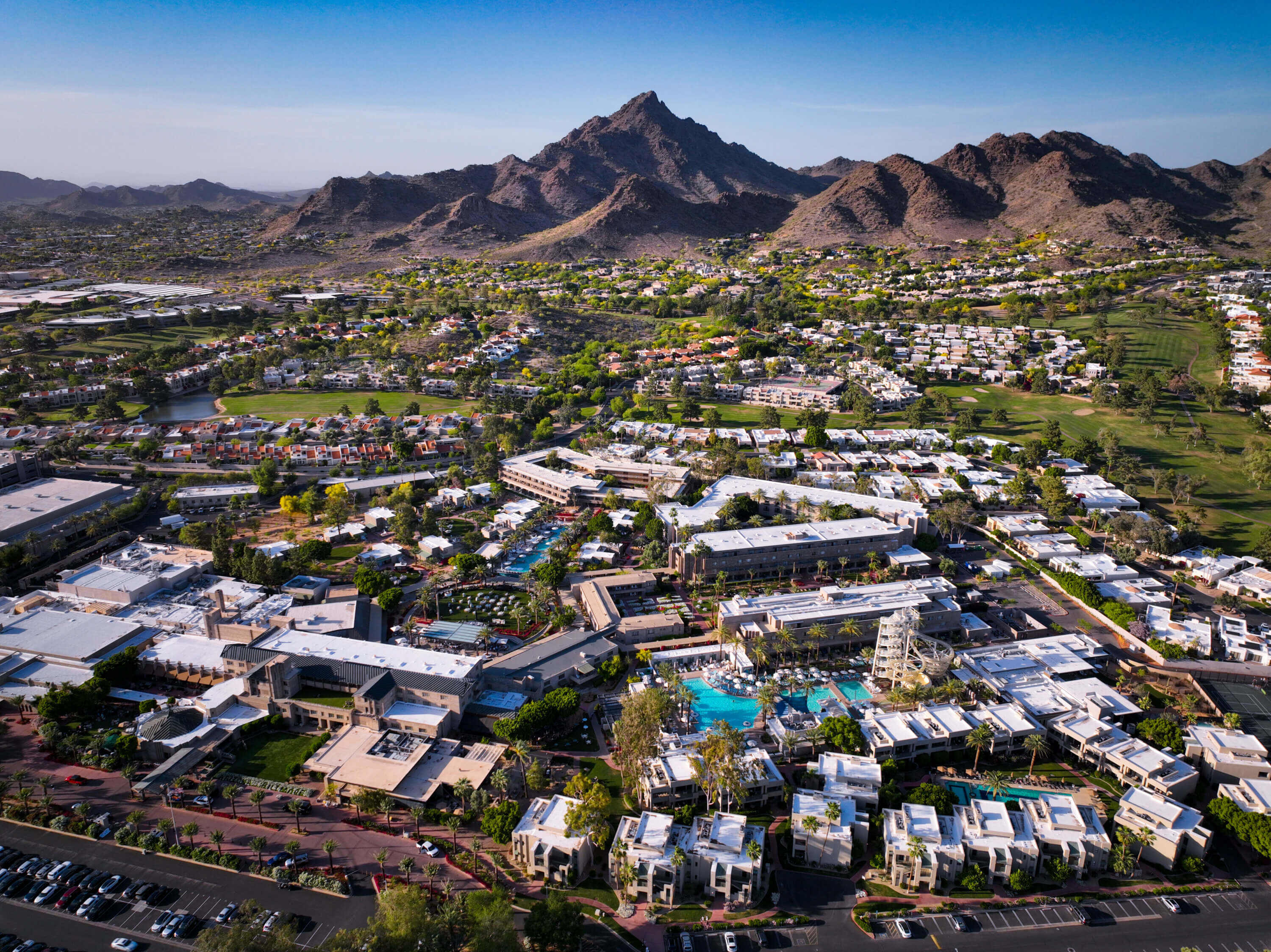 Aerial view of a resort complex with pools and white buildings, surrounded by lush landscaping and palm trees—your Championship Weekend Escape set against rugged mountains and a sprawling suburb under a clear blue sky.