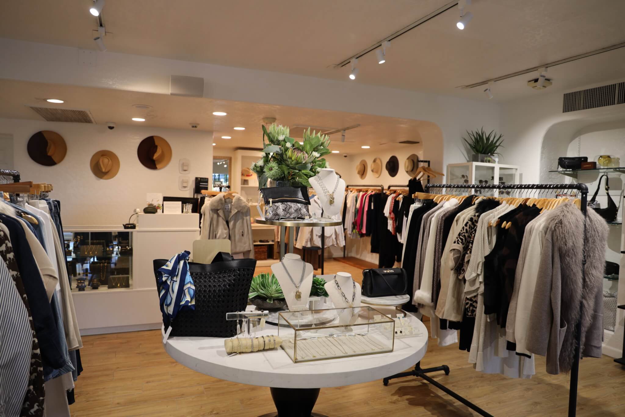 A stylish boutique with racks of clothing, handbags, and accessories. A round table in front displays jewelry, bags, and a plant. The space is well-lit with wooden floors and hats decoratively mounted on the walls.