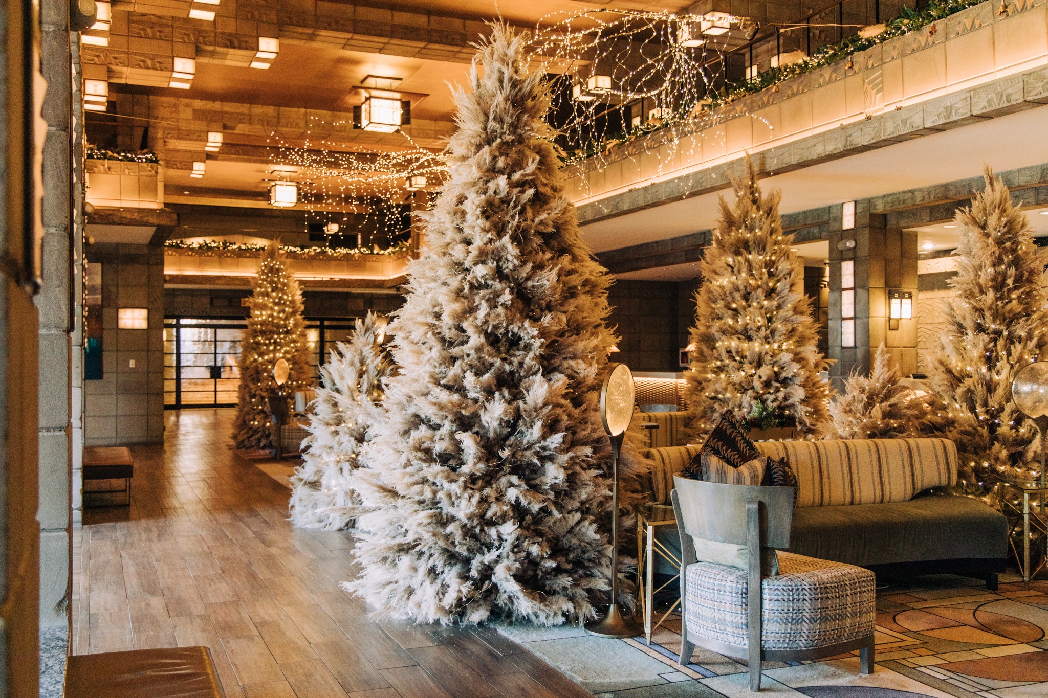 A spacious, elegant lobby decorated with large, snow-frosted Christmas trees, string lights, and festive greenery celebrates the holidays at the Arizona Biltmore. Cozy seating areas with modern chairs and a sofa are arranged on a wooden floor.