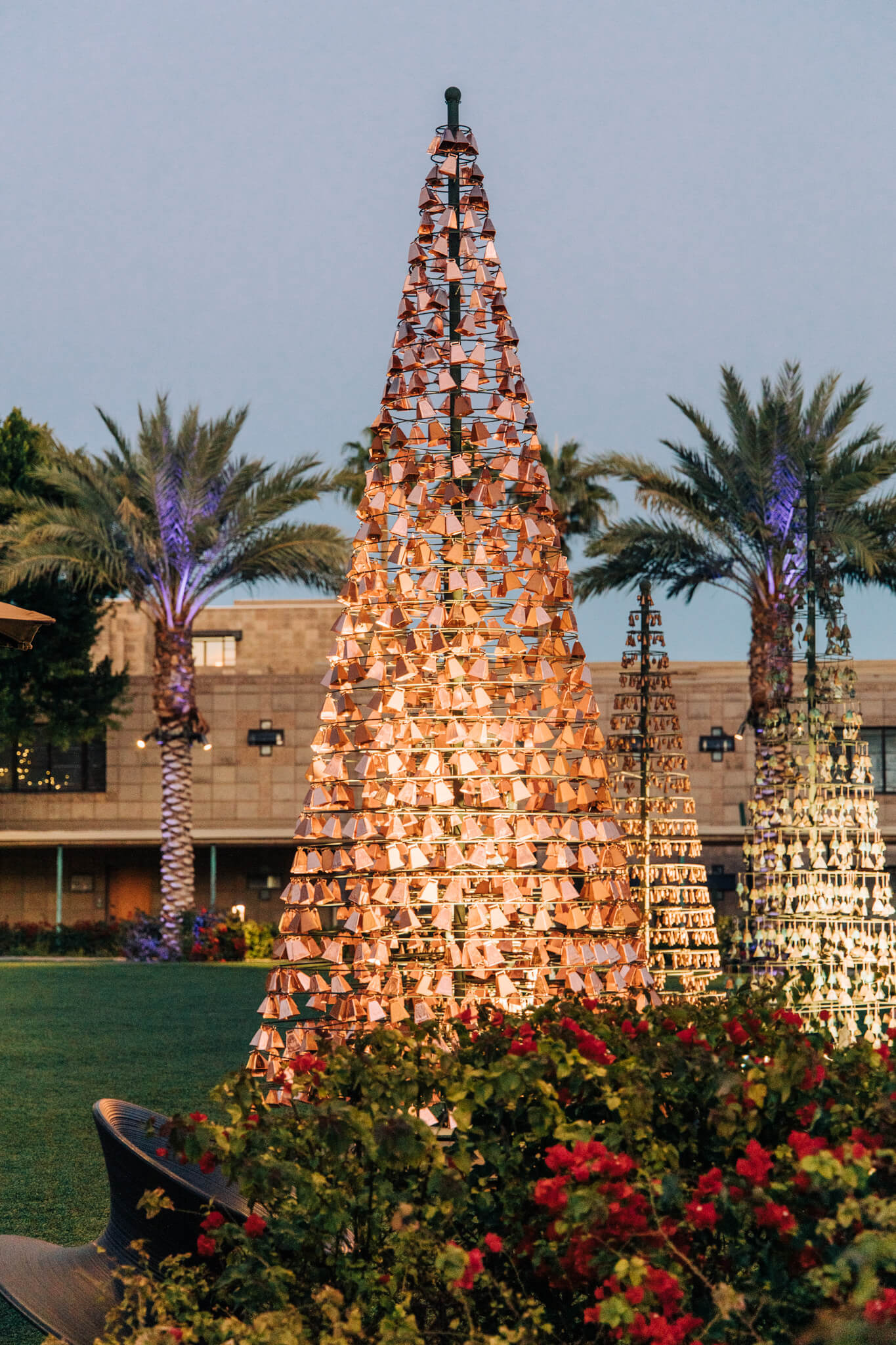 Decorative, illuminated metal cone sculptures resembling Christmas trees—some even shaped like a taco—are displayed outdoors among flowers and palm trees at dusk.