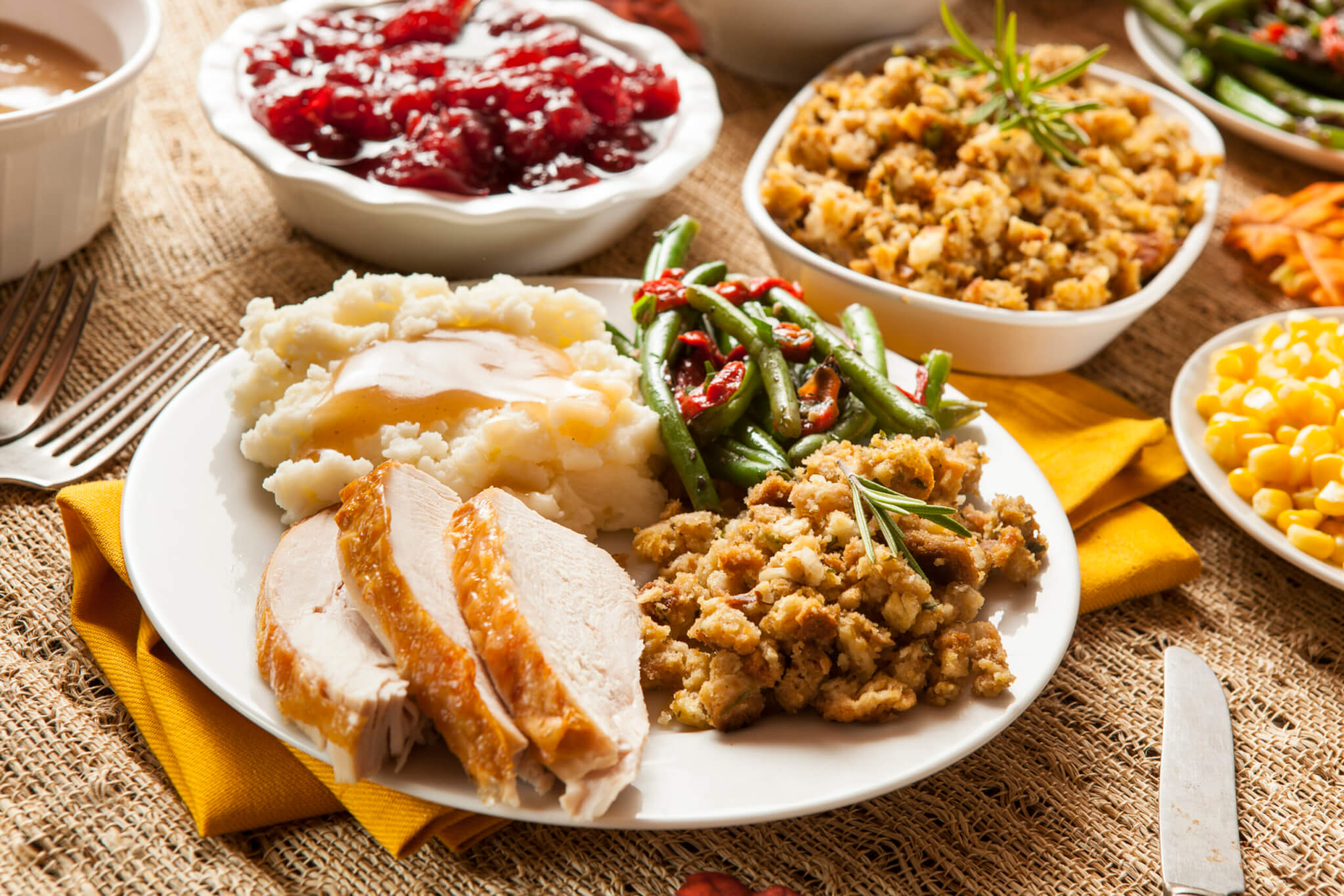 A plate of Thanksgiving food with sliced turkey, mashed potatoes and gravy, green beans, stuffing, and a side of corn. A festive table holds more dishes like cranberry sauce—and even a turkey burger for a modern twist—ready to enjoy.