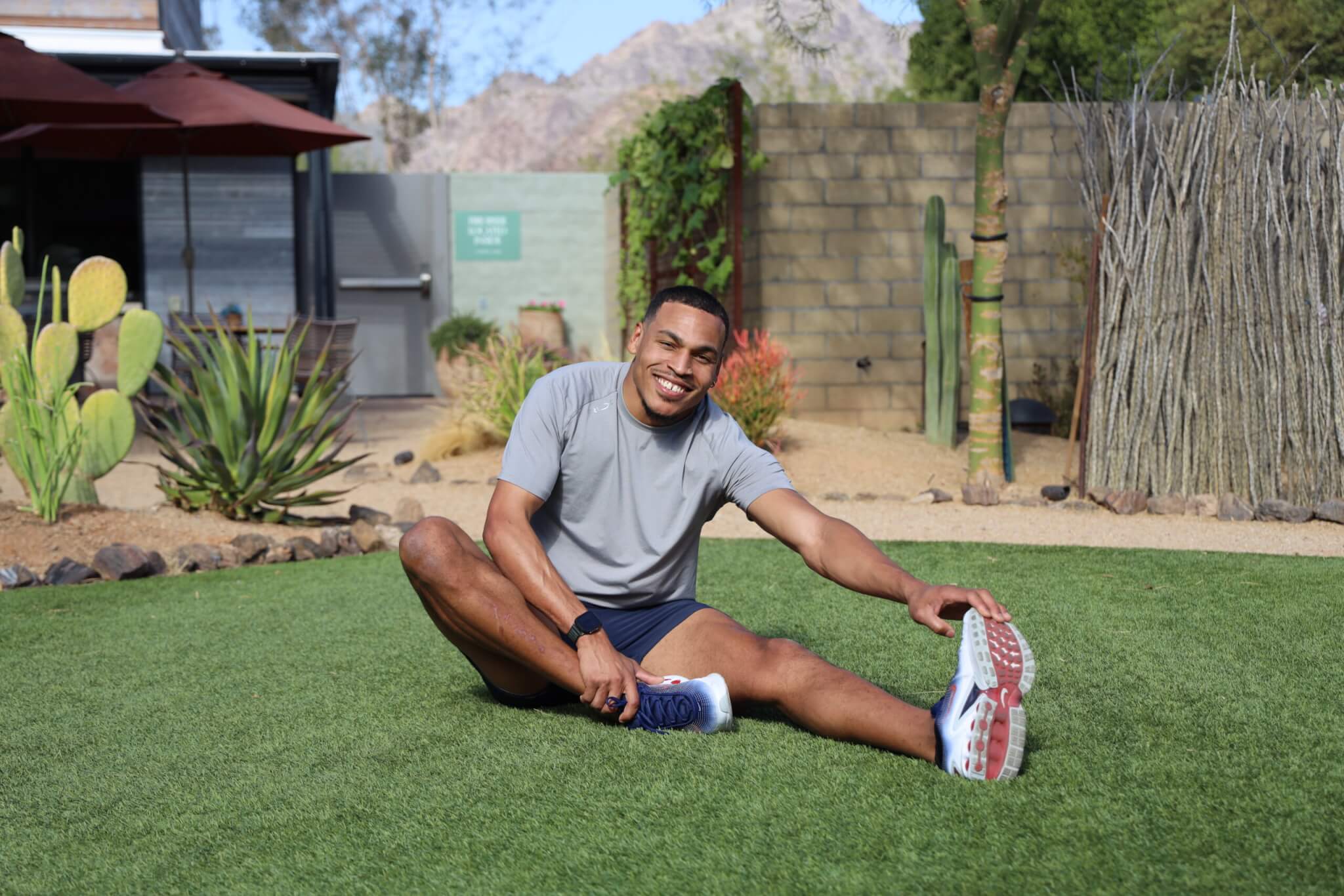 A man wearing a gray t-shirt and shorts sits on green grass, smiling while stretching his legs outdoors in a garden with cacti, plants, and a stone wall in the background.