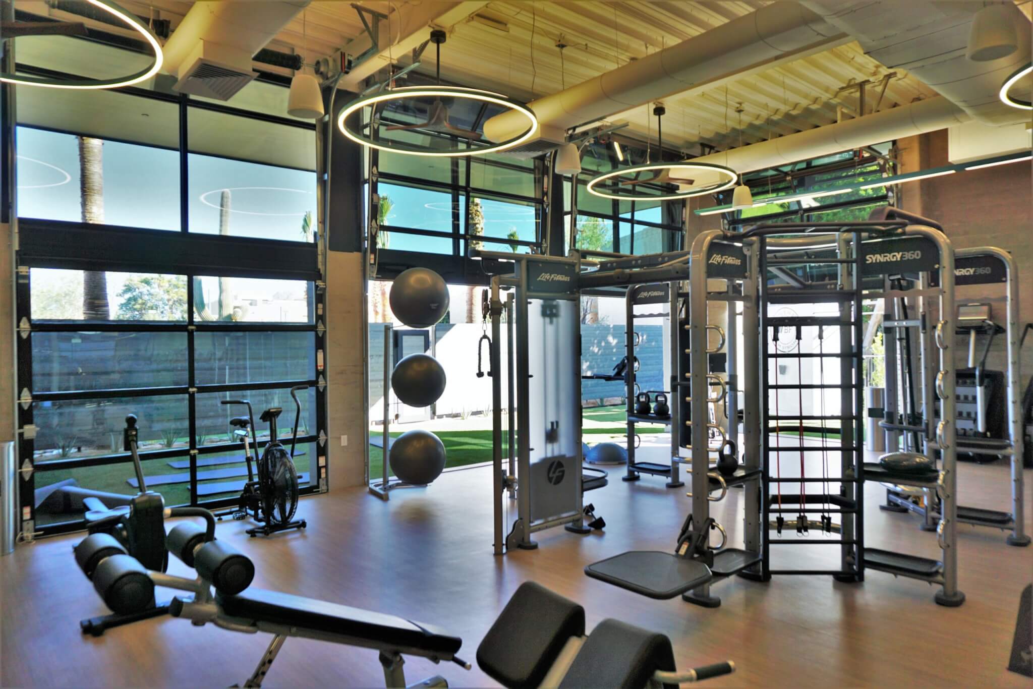 Modern gym with large windows, exercise machines, weight benches, medicine balls, and bright ring lights overhead. Natural light fills the spacious room, and outdoor greenery is visible through the windows.