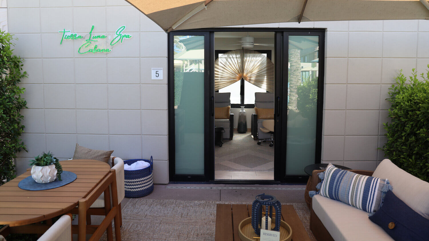 Outdoor patio area with a round wooden table, cushioned seating, and greenery. Glass doors open to a modern interior spa with chairs and a large decorative fan. Ask about our NYE Package at Terese Lane Spa Cabana, lit by a neon sign on the left wall.