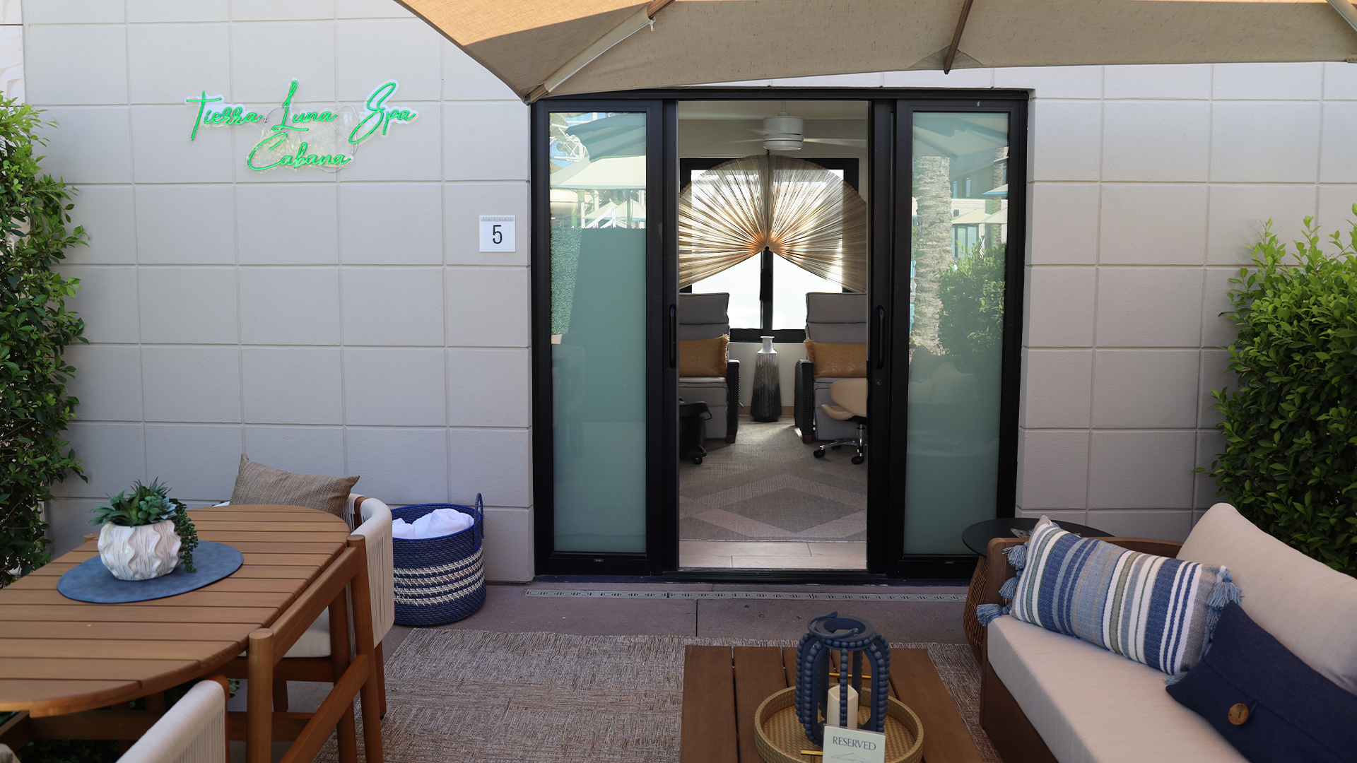 Outdoor patio area with a round wooden table, cushioned seating, and greenery. Glass doors open to a modern interior spa with chairs and a large decorative fan. Ask about our NYE Package at Terese Lane Spa Cabana, lit by a neon sign on the left wall.