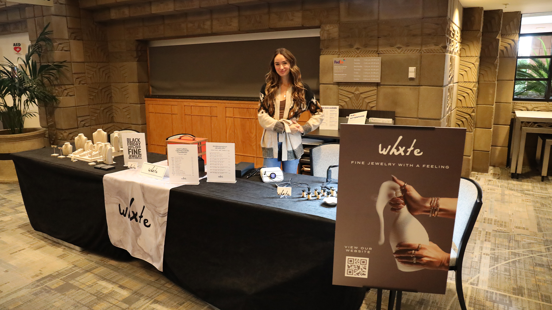A woman stands behind a display table featuring jewelry and promotional materials for Wxte, a fine jewelry brand, at an indoor event. The table has a white cloth, signs, and jewelry pieces on display.