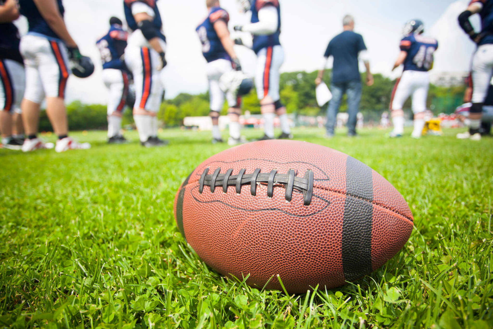 A close-up of an American football lying on green grass during spring training, with a group of players and a coach standing in the background, slightly out of focus.