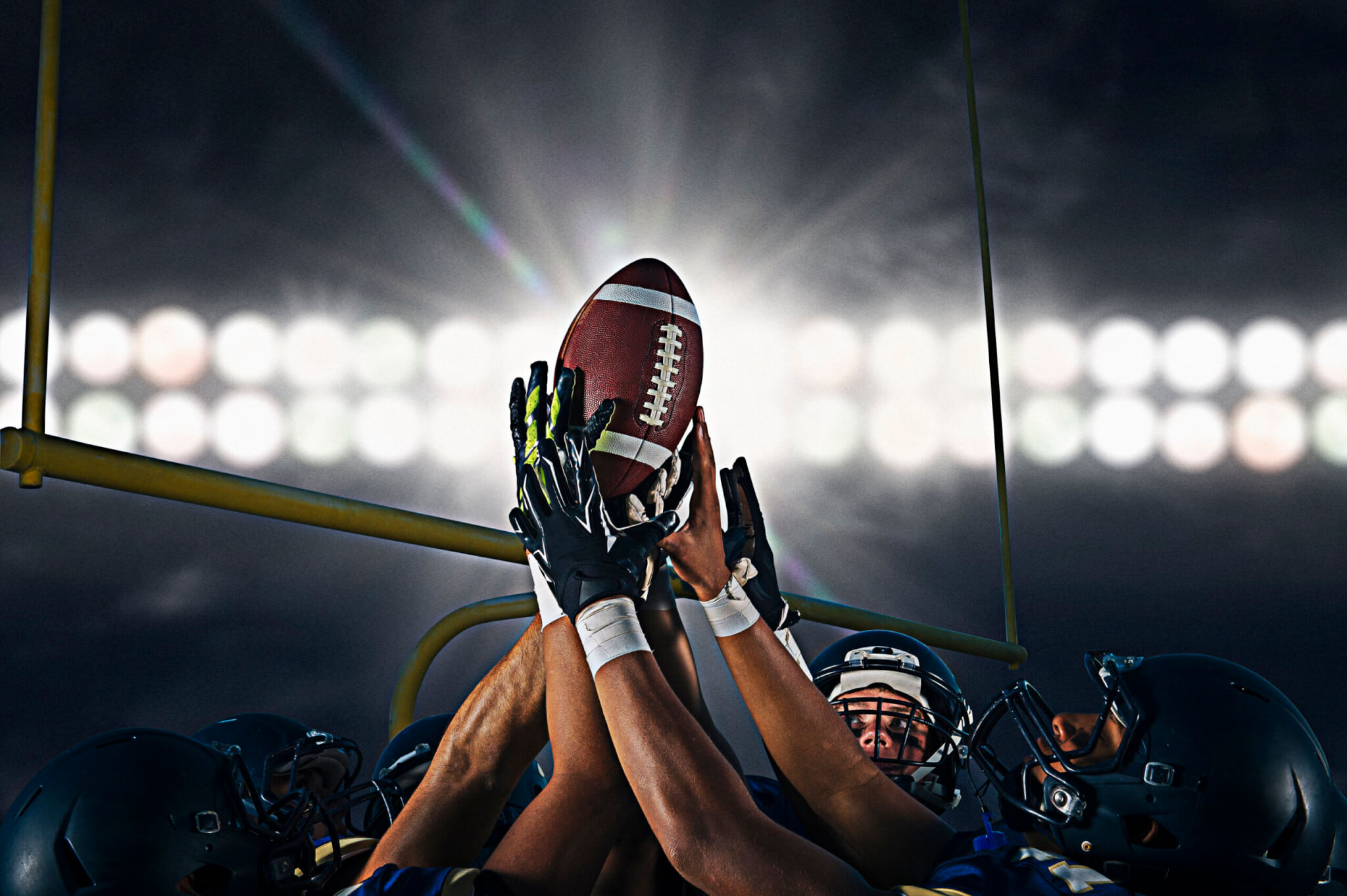 A group of American football players, wearing helmets and gloves, reach up together for a football near the goalpost under bright stadium lights at night, reminiscent of the excitement found during holidays at the Arizona Biltmore.