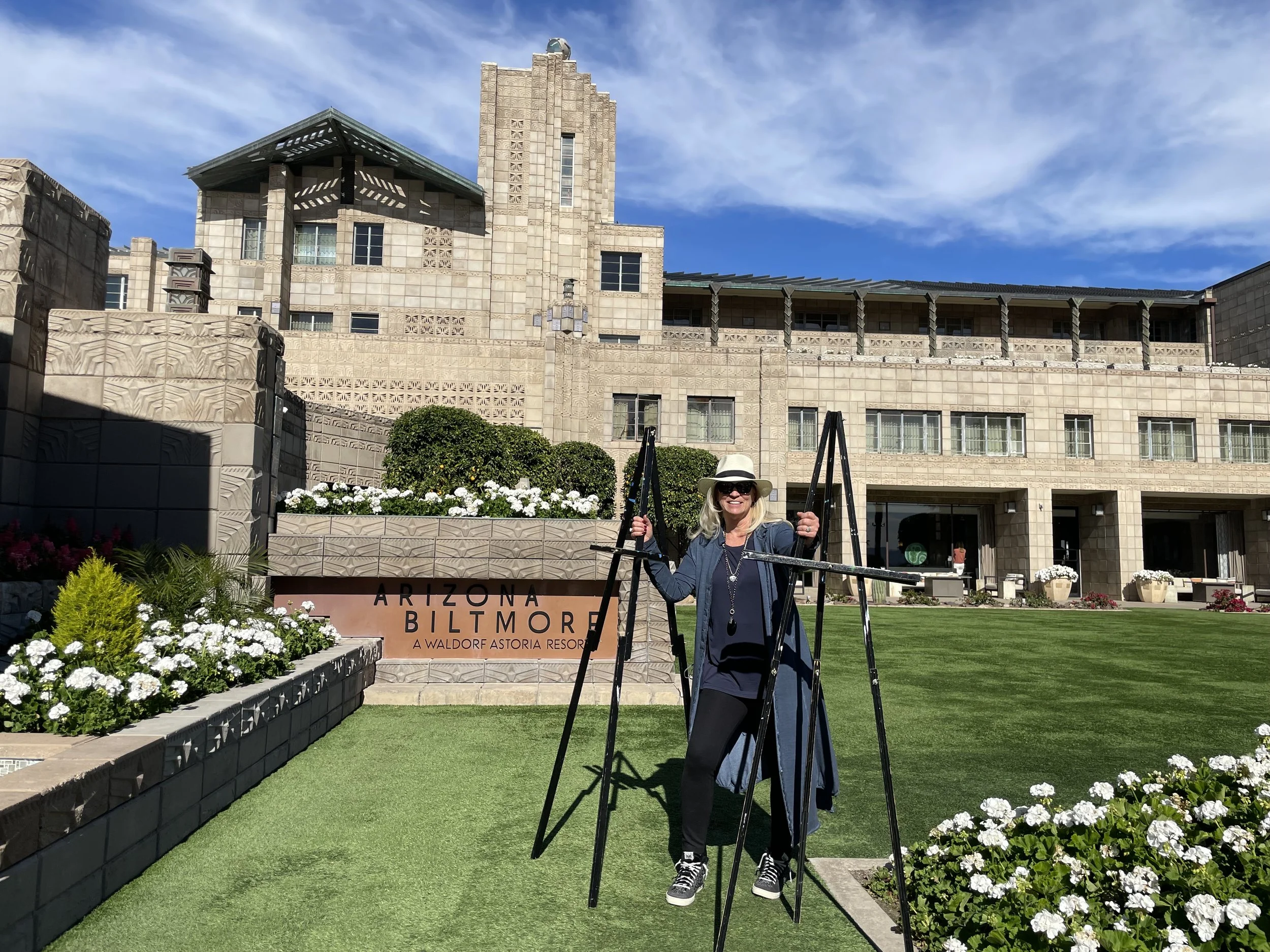 A woman in sunglasses and a hat stands on stilts on a grassy lawn in front of the Arizona Biltmore resort, where Gingerbread House Decorating events capture the festive spirit amid blue skies and stunning architecture.