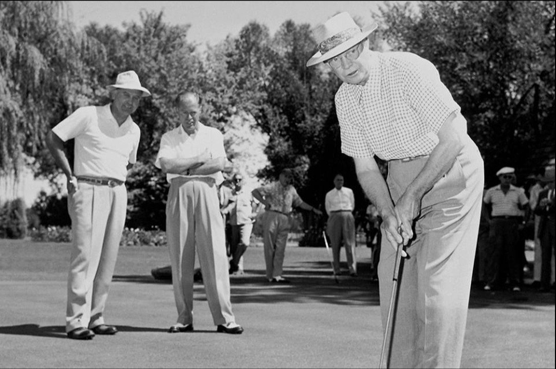 An older man in a hat and checkered shirt lines up a putt on mcarthur's golf course while several men in casual clothes and hats watch him play on a sunny day, with trees visible in the background.