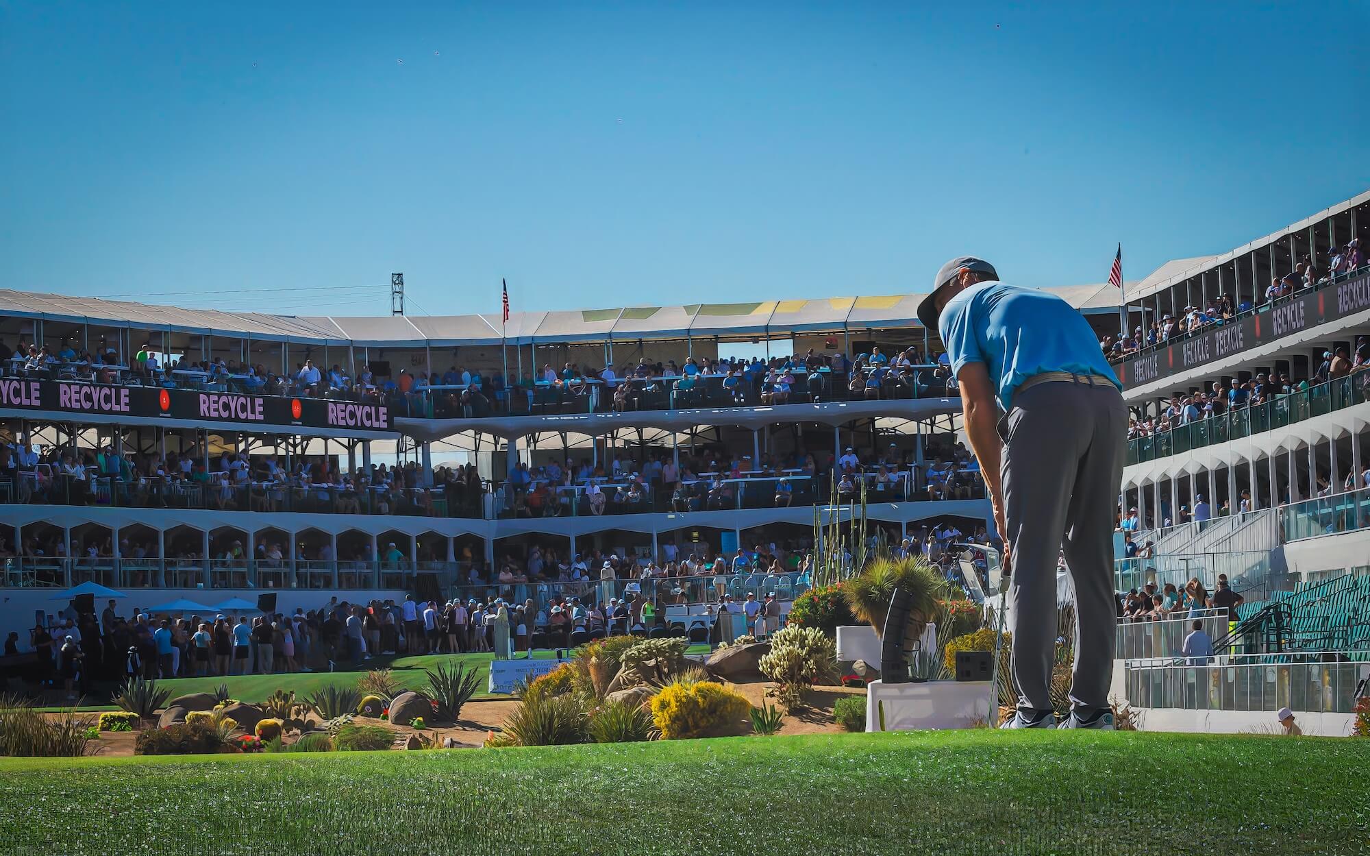 A golfer in blue prepares to putt on a green, while a large crowd watches from multi-level grandstands under a clear blue sky at a professional golf tournament during spring training.