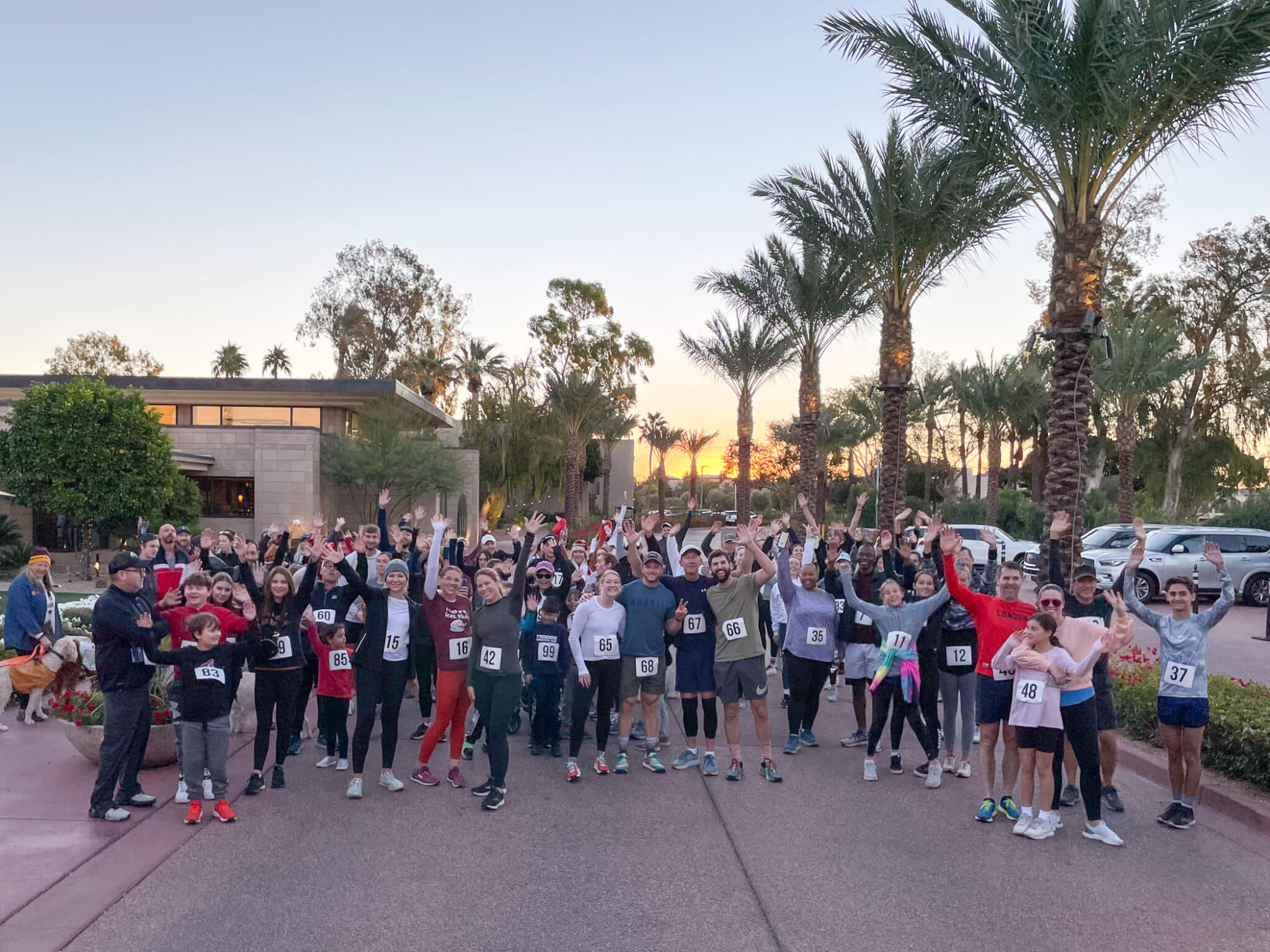 A large group of people, many wearing numbered race bibs, pose and cheer together outdoors near palm trees at sunrise or sunset, suggesting the start or end of a fun run or community race.