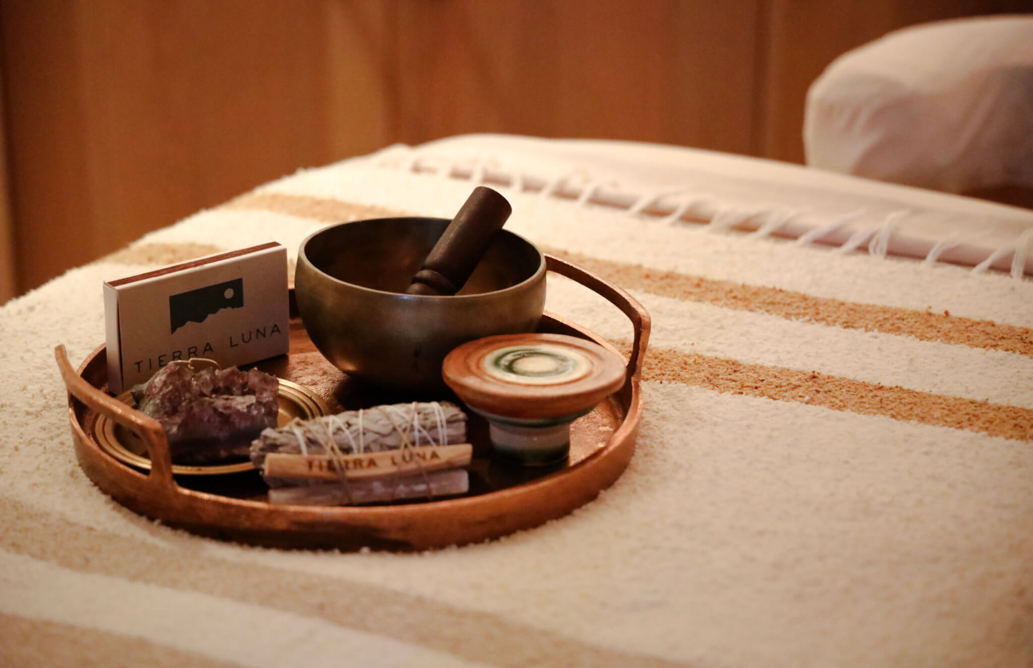 A wooden tray on a striped towel holds a singing bowl with mallet, a crystal, sage bundle, ceramic dish, and Tierra Luna cards—perfect for Full Moon or New Moon rituals in a serene, spa-like or meditation setting.