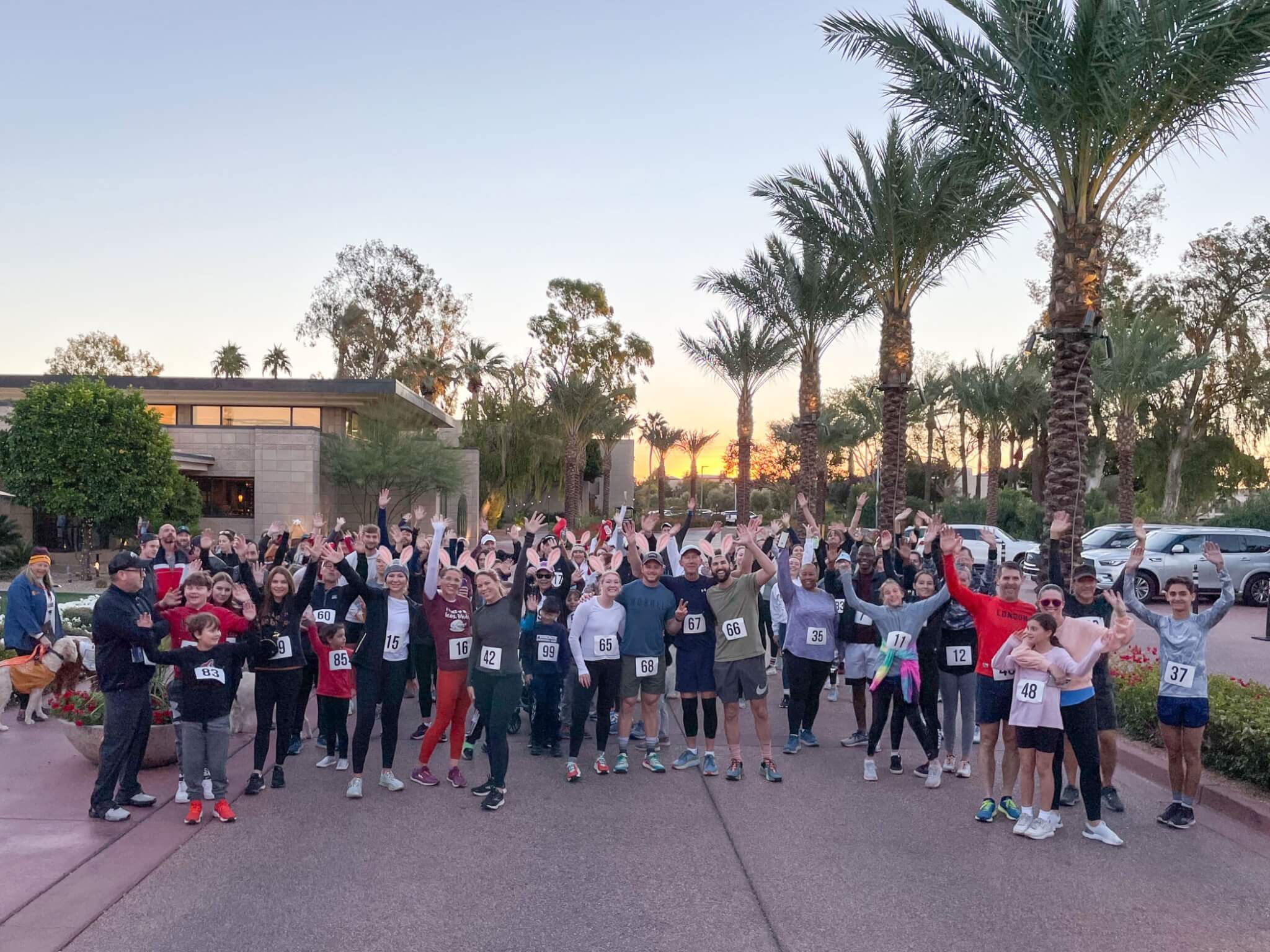 A large group of people wearing race bibs gather outdoors among palm trees at sunset, smiling and raising their arms, ready for a community fun run or race event.