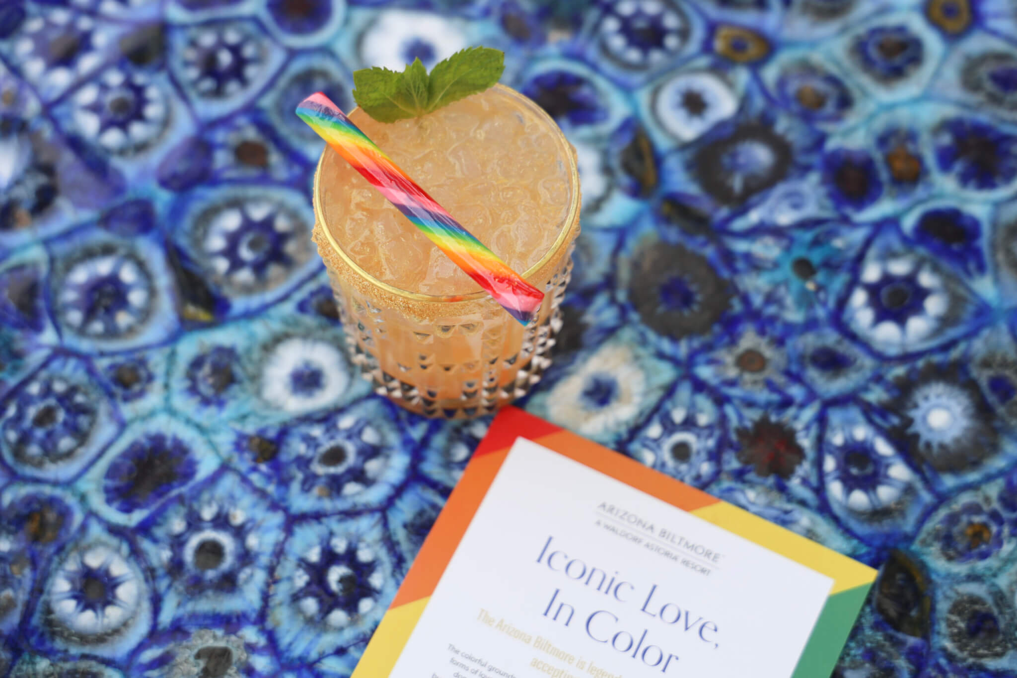 A colorful drink with a rainbow straw and mint garnish sits on a blue patterned surface next to a paper titled "Iconic Love: In Color" with a vibrant rainbow border, celebrating the spirit of Iconic Love.