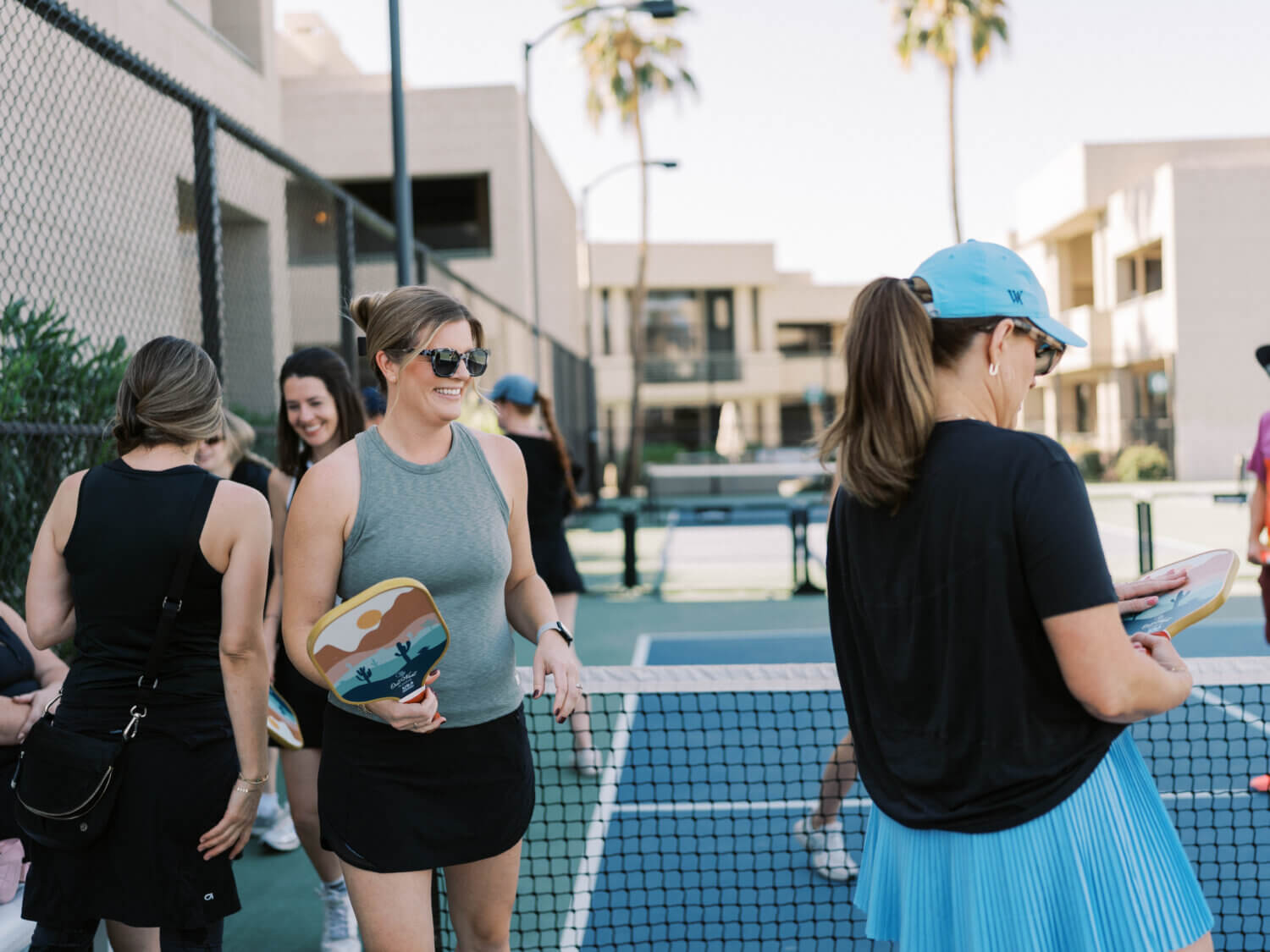 A group of women stand by a pickleball net on an outdoor court, smiling and talking on a warm summer day. One woman holds a paddle and wears sunglasses; another wears a blue skirt and cap. Palm trees and buildings are in the background.