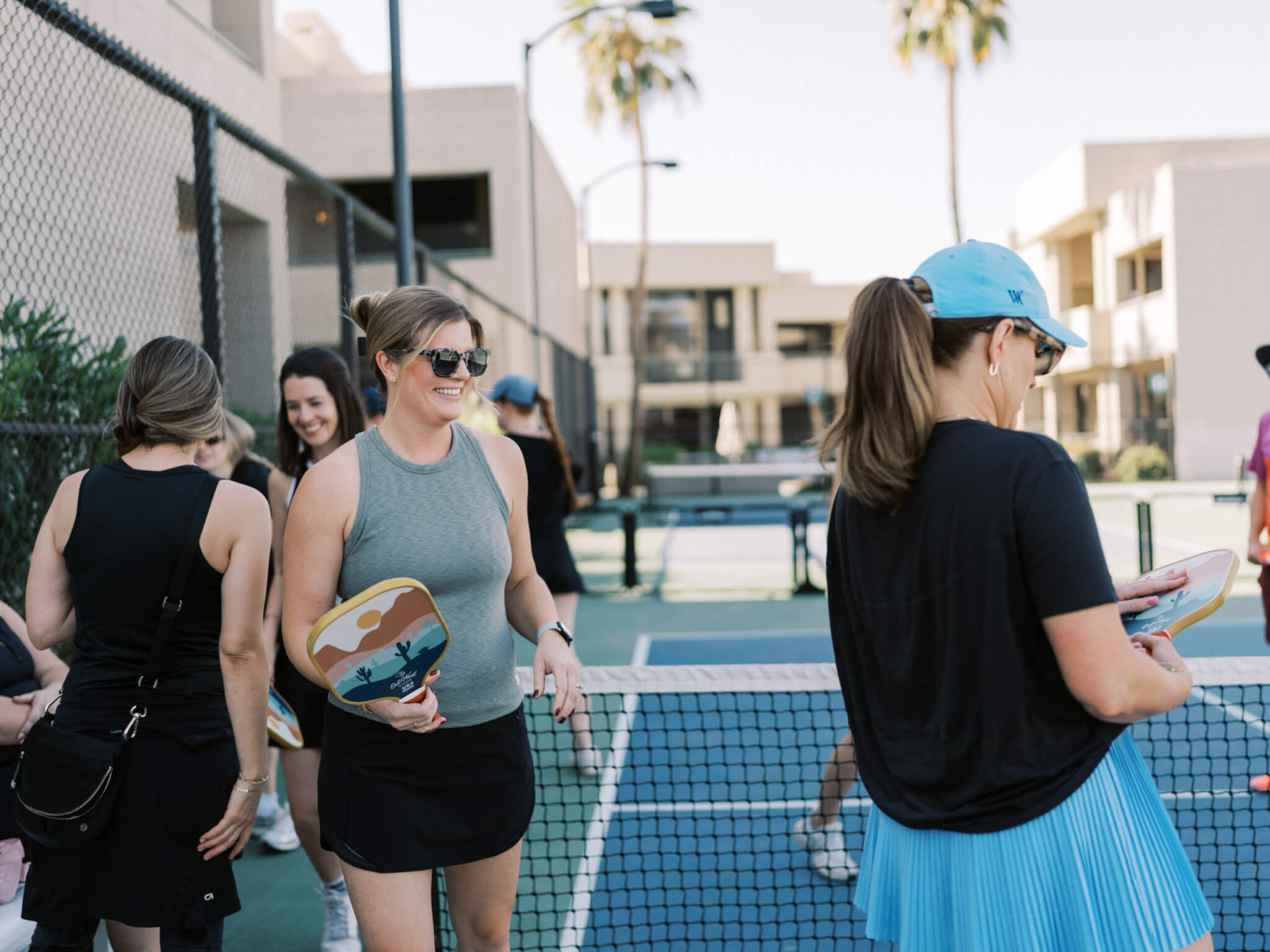 A group of women stand by a pickleball net on an outdoor court, smiling and talking on a warm summer day. One woman holds a paddle and wears sunglasses; another wears a blue skirt and cap. Palm trees and buildings are in the background.