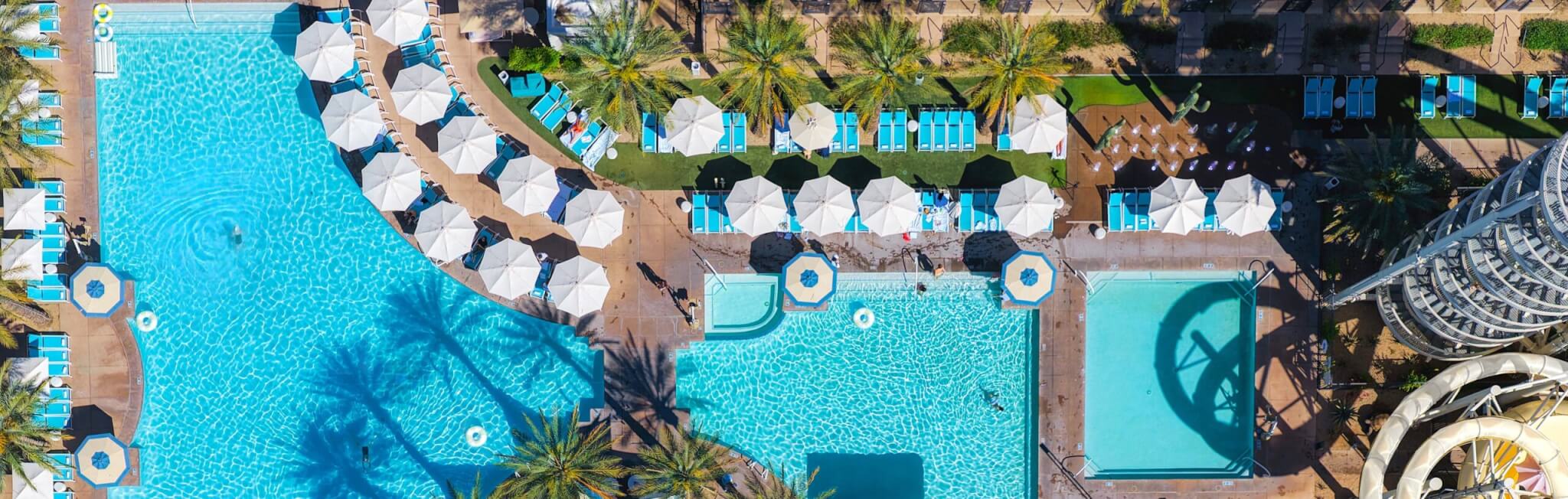 Aerial view of the Arizona Biltmore’s large outdoor swimming pool area with blue water, white umbrellas, lounge chairs, palm trees, and water slides under bright sunlight.