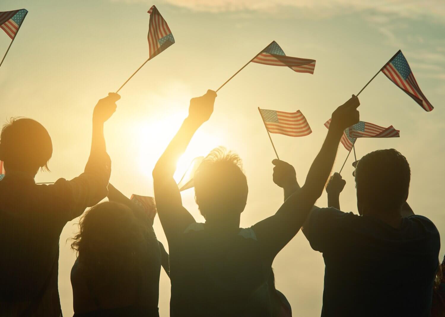 Silhouettes of people holding and waving small American flags in the air, with the sun shining brightly behind them. Nearby, a bike rental station adds to the festive atmosphere, suggesting a lively patriotic celebration.