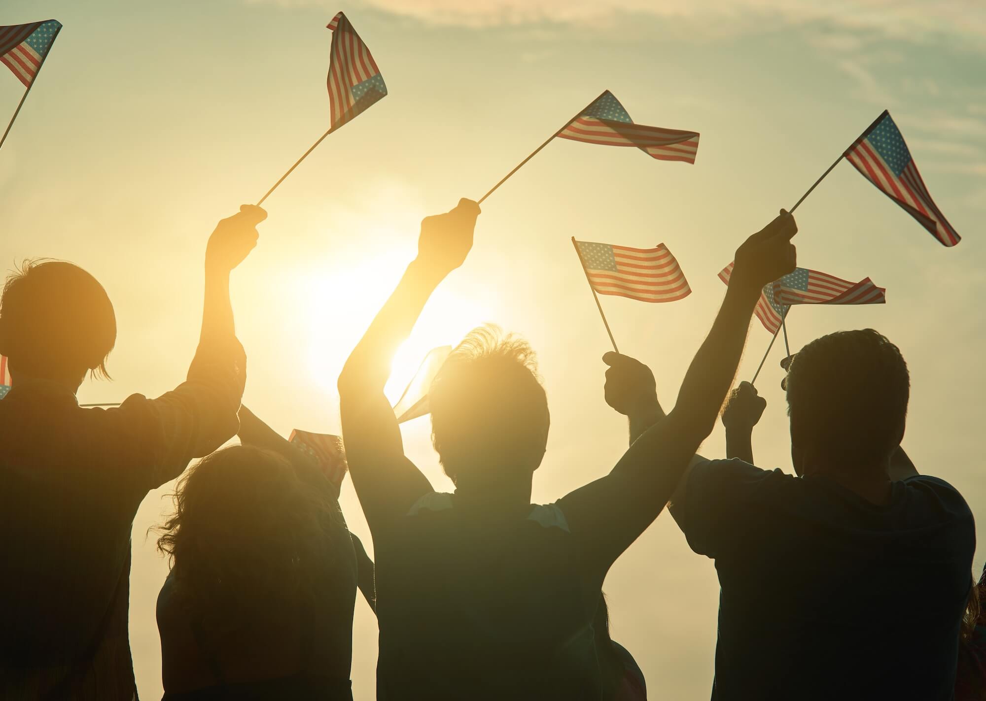 Silhouettes of people holding and waving small American flags in the air, with the sun shining brightly behind them. Nearby, a bike rental station adds to the festive atmosphere, suggesting a lively patriotic celebration.