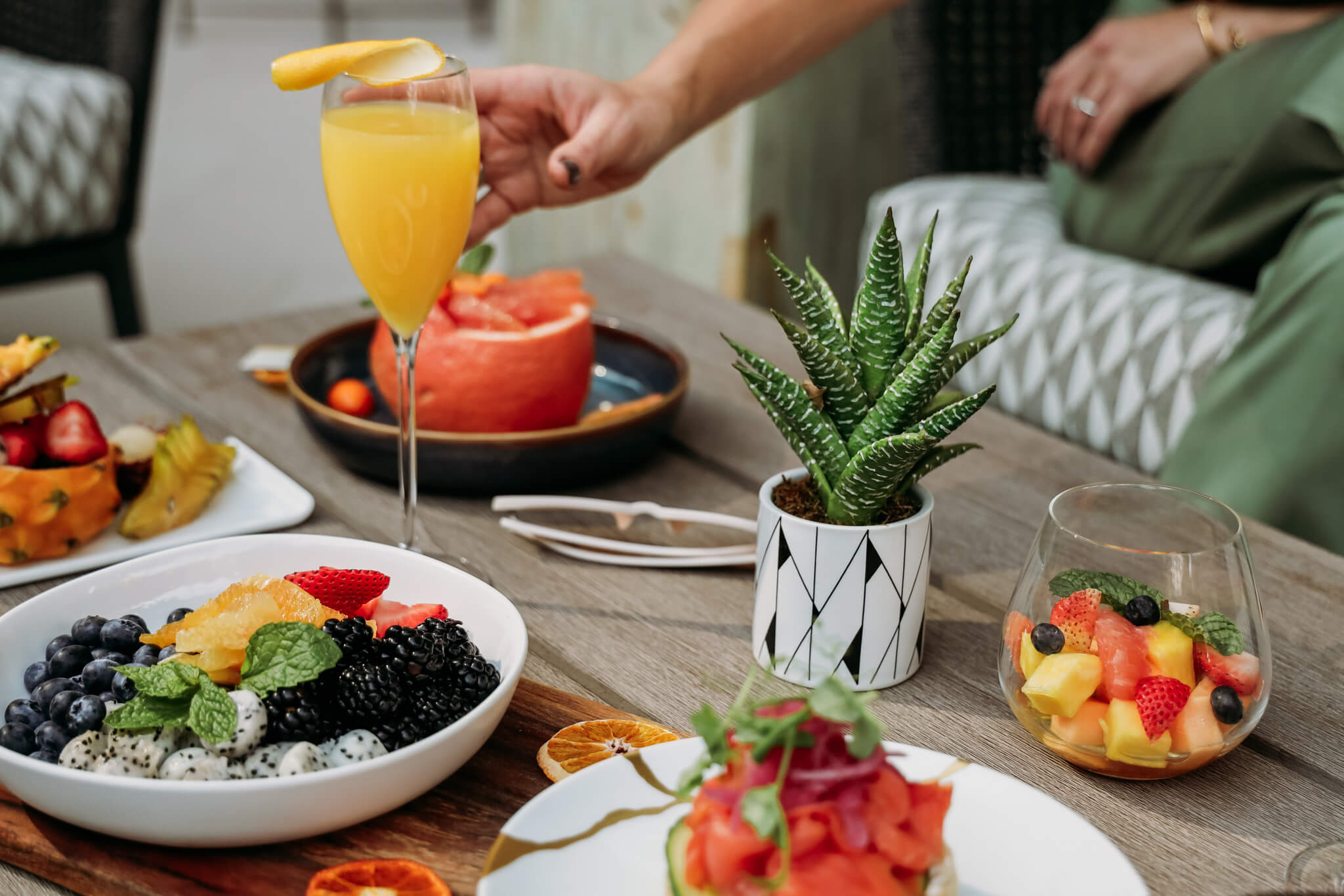 A brunch table with fresh fruit bowls, a glass of orange juice with a slice on the rim, a small potted succulent, and a person reaching for food. Plates include berries, watermelon, and assorted colorful fruit.