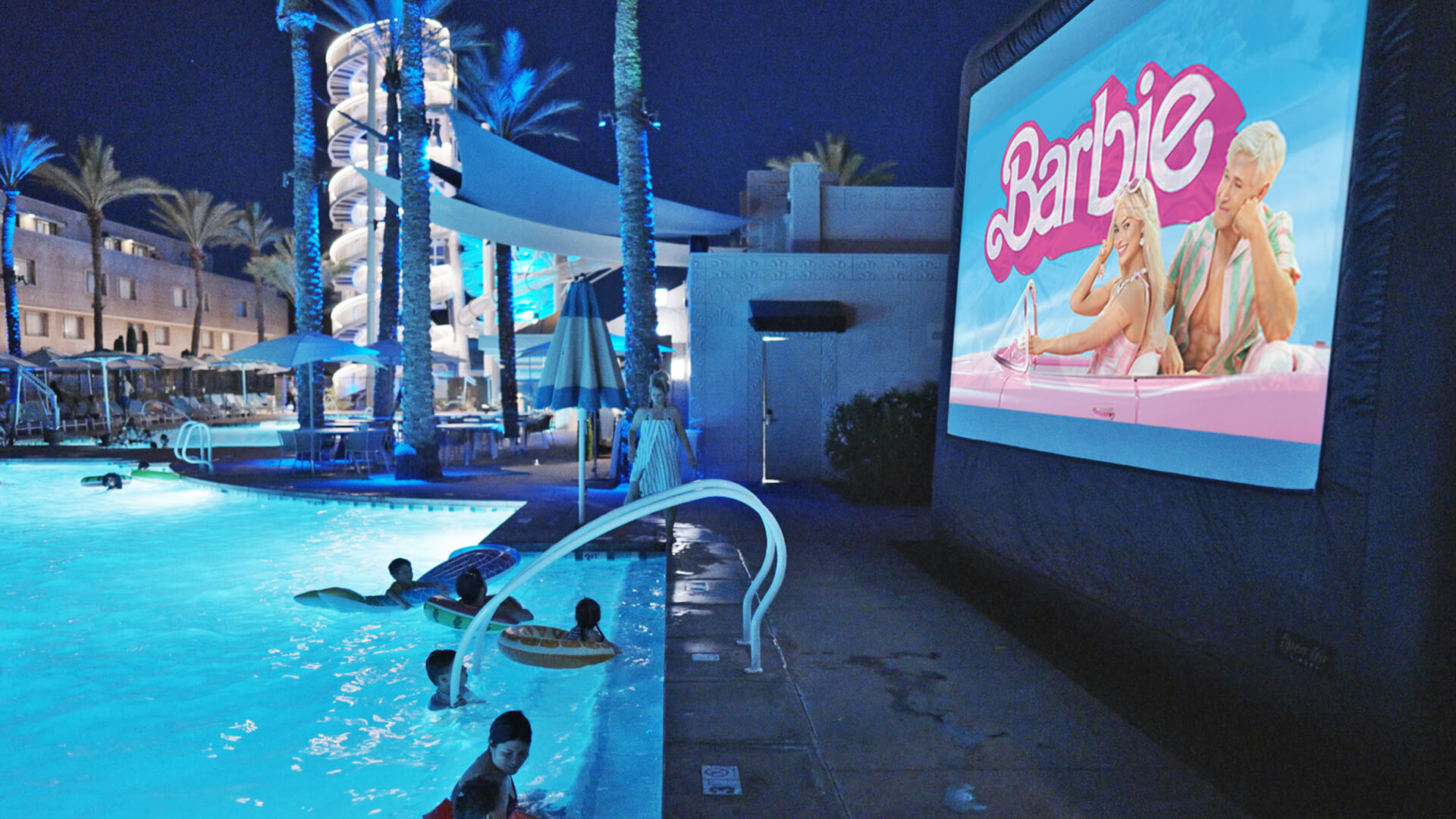 People swim in a pool at night while an outdoor movie screen shows the Barbie movie. Palm trees, umbrellas, and buildings with lights are visible in the background.