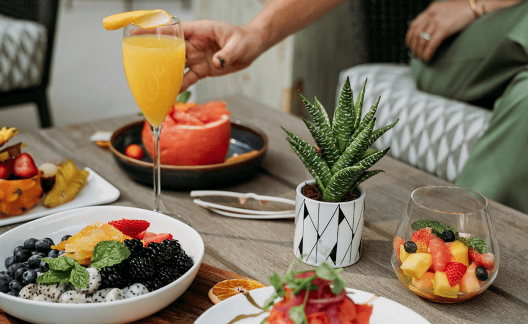 A hand reaches for a glass of orange juice on a wooden table set with bowls of fresh fruit, a small potted succulent, and a glass of mixed fruit. Another person sits in the background.